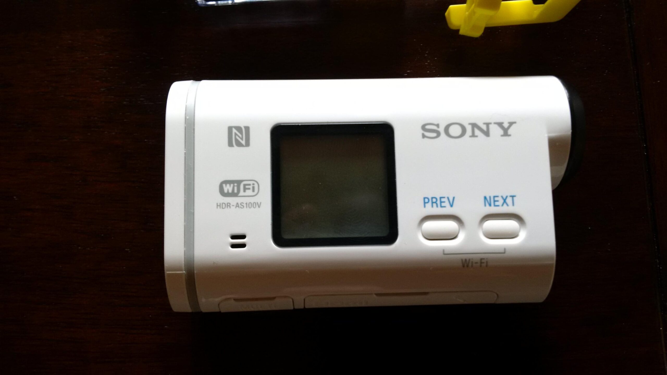 Sony HDR-AS100VR: A top-down view of a white Sony action camera featuring a small display screen, control buttons labeled "PREV" and "NEXT," and icons for NFC and Wi-Fi connectivity. The model number "HDR-AS100V" is visible on the device. The camera is placed on a dark wooden surface, with a yellow plastic clip partially visible in the background.