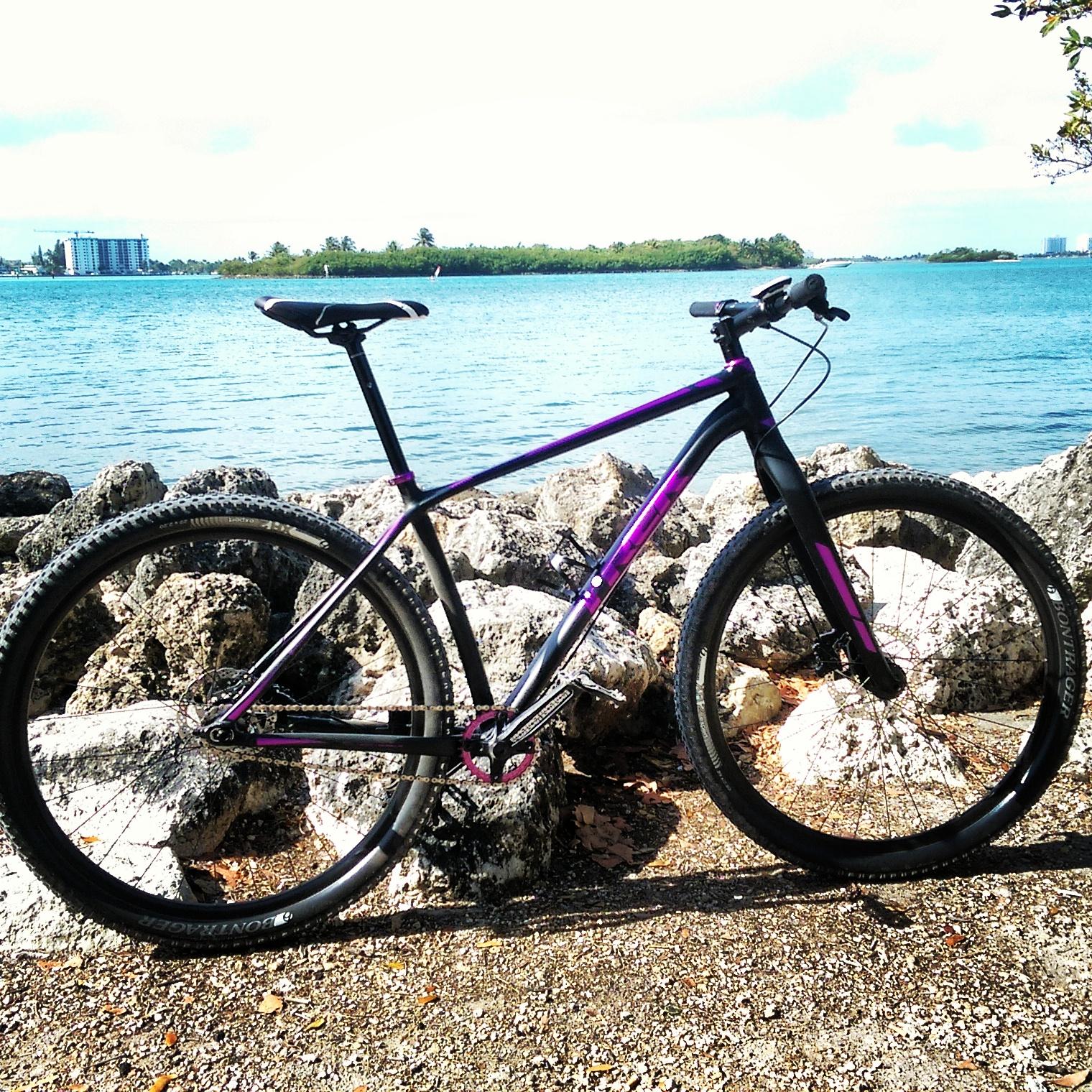 A mountain bike with a black and purple frame is positioned on rocky terrain by a body of water, under a bright blue sky with a few clouds. In the background, a building and lush green foliage can be seen along the water's edge. Oleta River State Park mountain bike trail.
