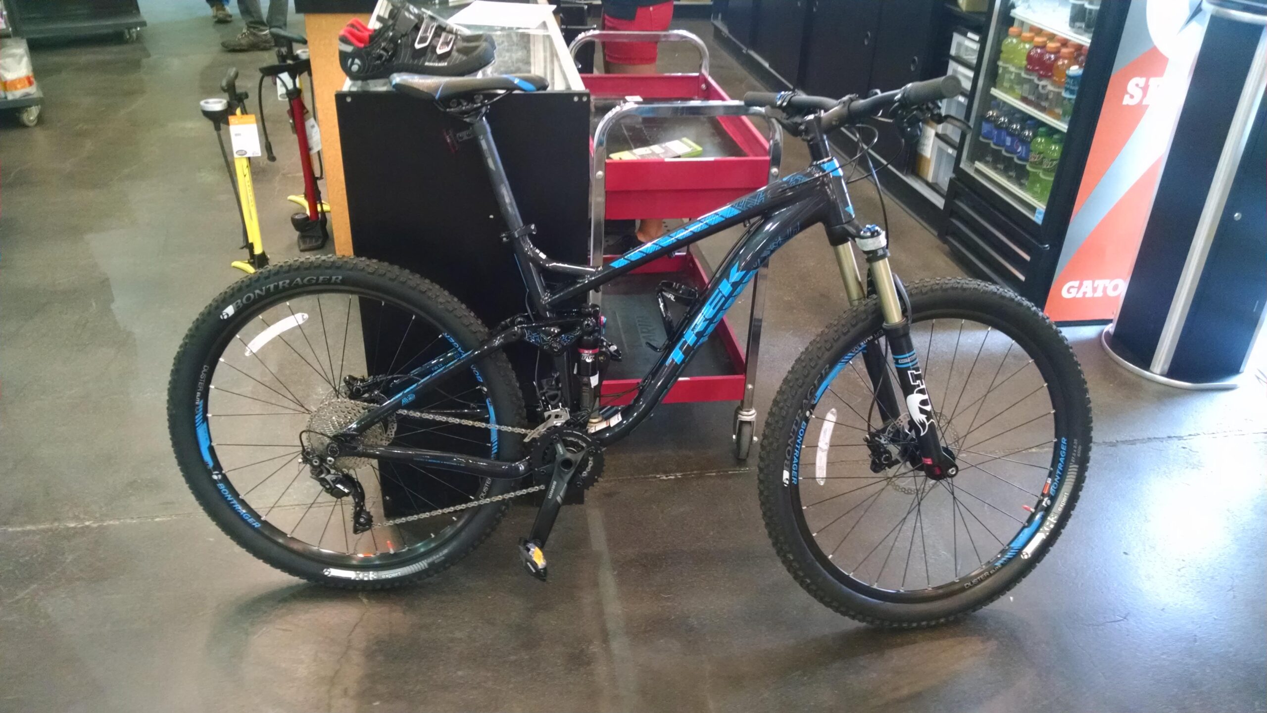 Trek Fuel EX 7: A black mountain bike with blue accents, featuring knobby tires, a front suspension fork, and a visible gear system. The bike is positioned in a retail shop, with a red cart in the background and various cycling accessories nearby. The floor is dark and polished.