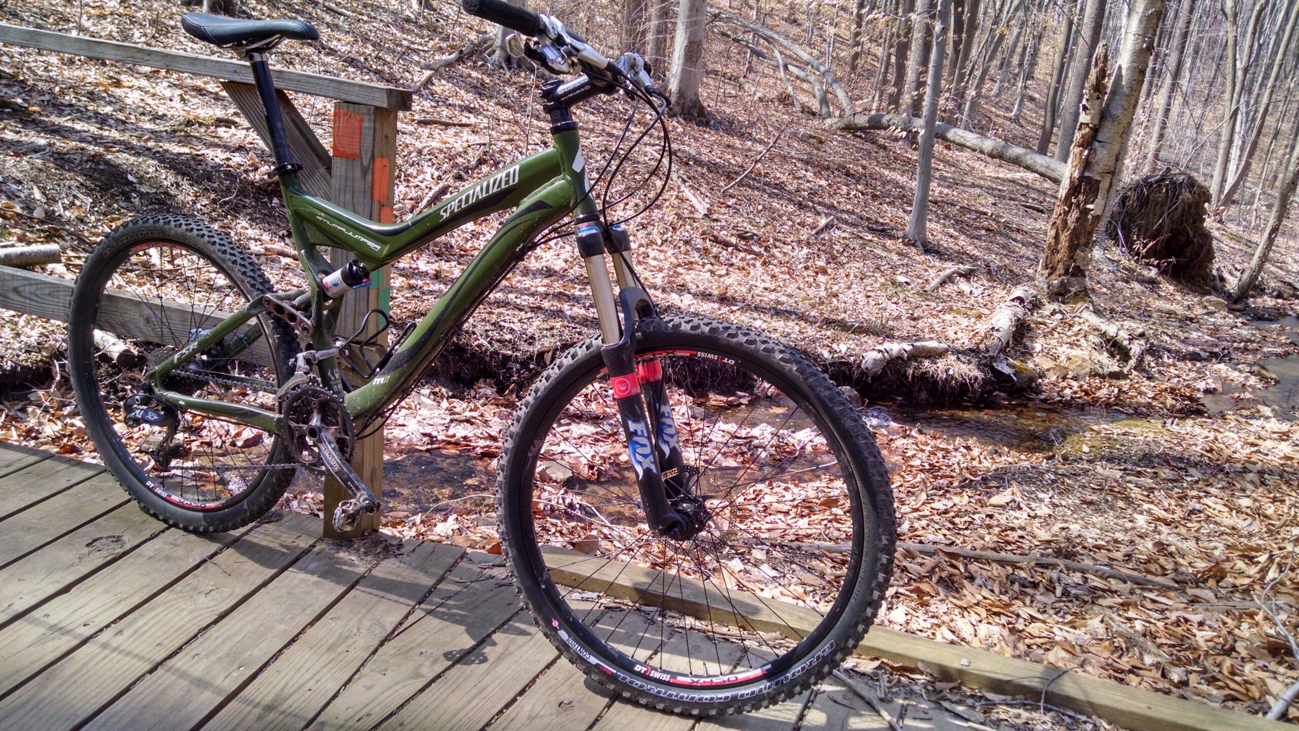 Specialized Stumpjumper FSR: A green mountain bike parked on a wooden platform in a forest. The background features leaf-covered ground, bare trees, and a small stream flowing beside the bike. The bike is equipped with a water bottle and features visible suspension components.