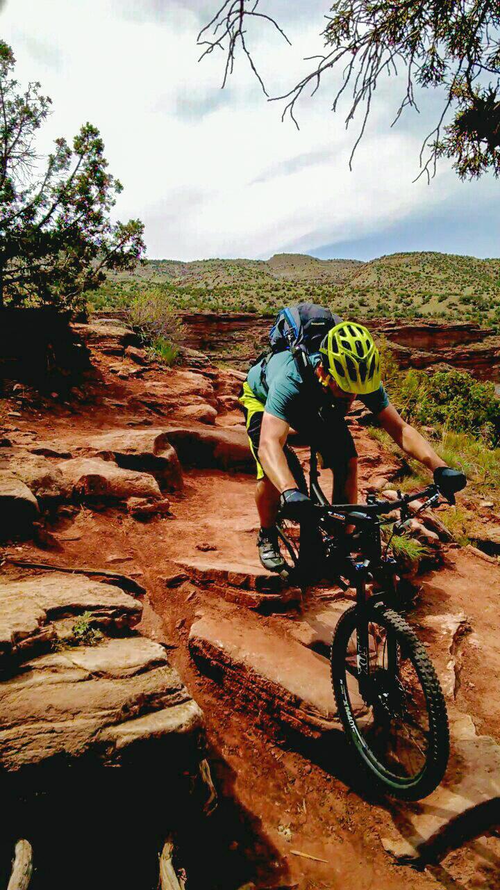 A mountain biker navigates a rocky trail in a rugged outdoor landscape, wearing a yellow helmet and a backpack. The terrain features reddish rocks and scattered vegetation under a cloudy sky. Mary's Loop / Horsethief Bench mountain bike trail.