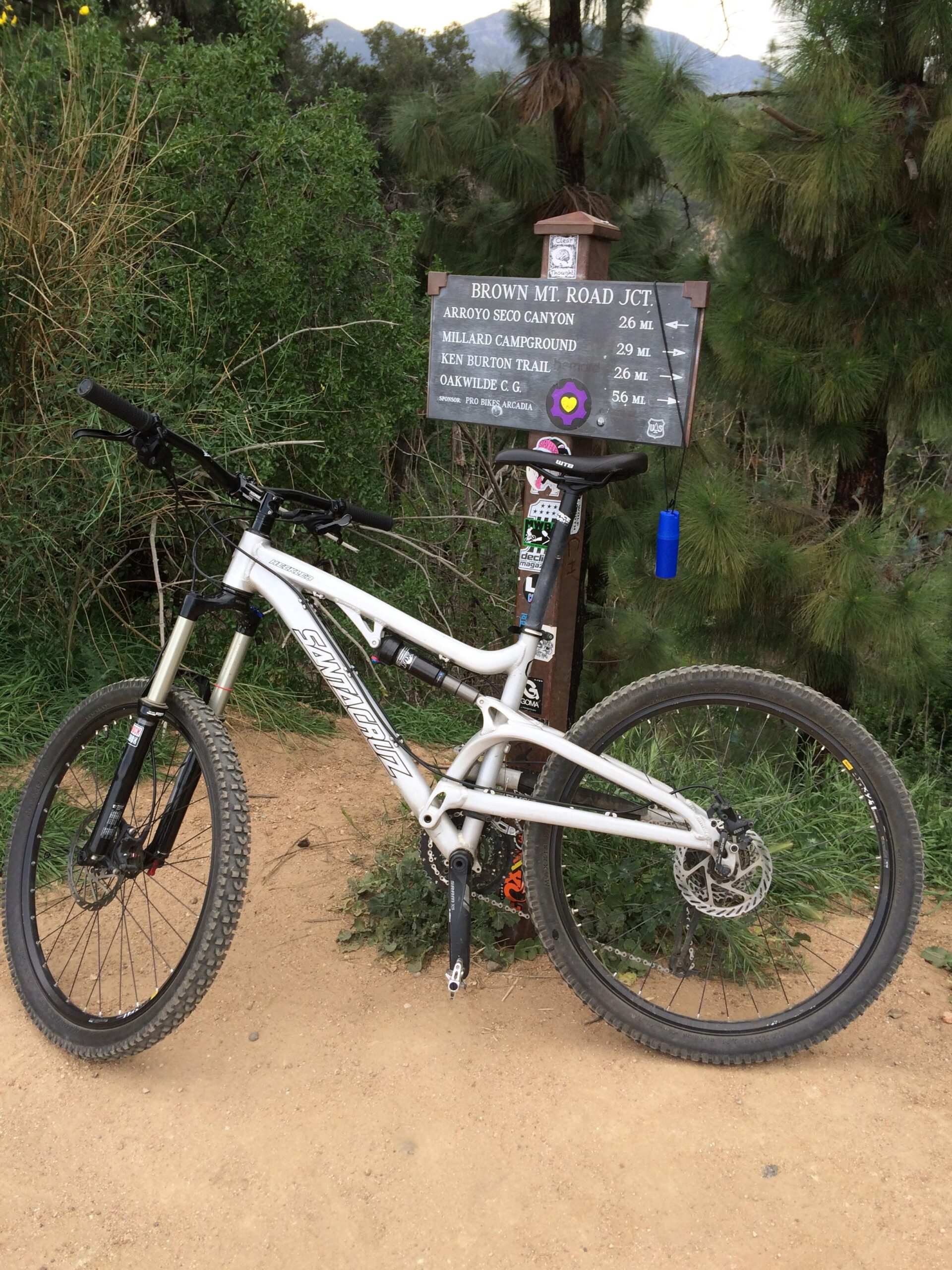 Santa Cruz Heckler: A mountain bike is parked next to a trail sign indicating directions and distances to various locations, including Arroyo Seco Canyon, Millard Campground, Ken Burton Trail, and Oakwilde Campground. The scene is set in a natural environment with greenery and trees in the background.