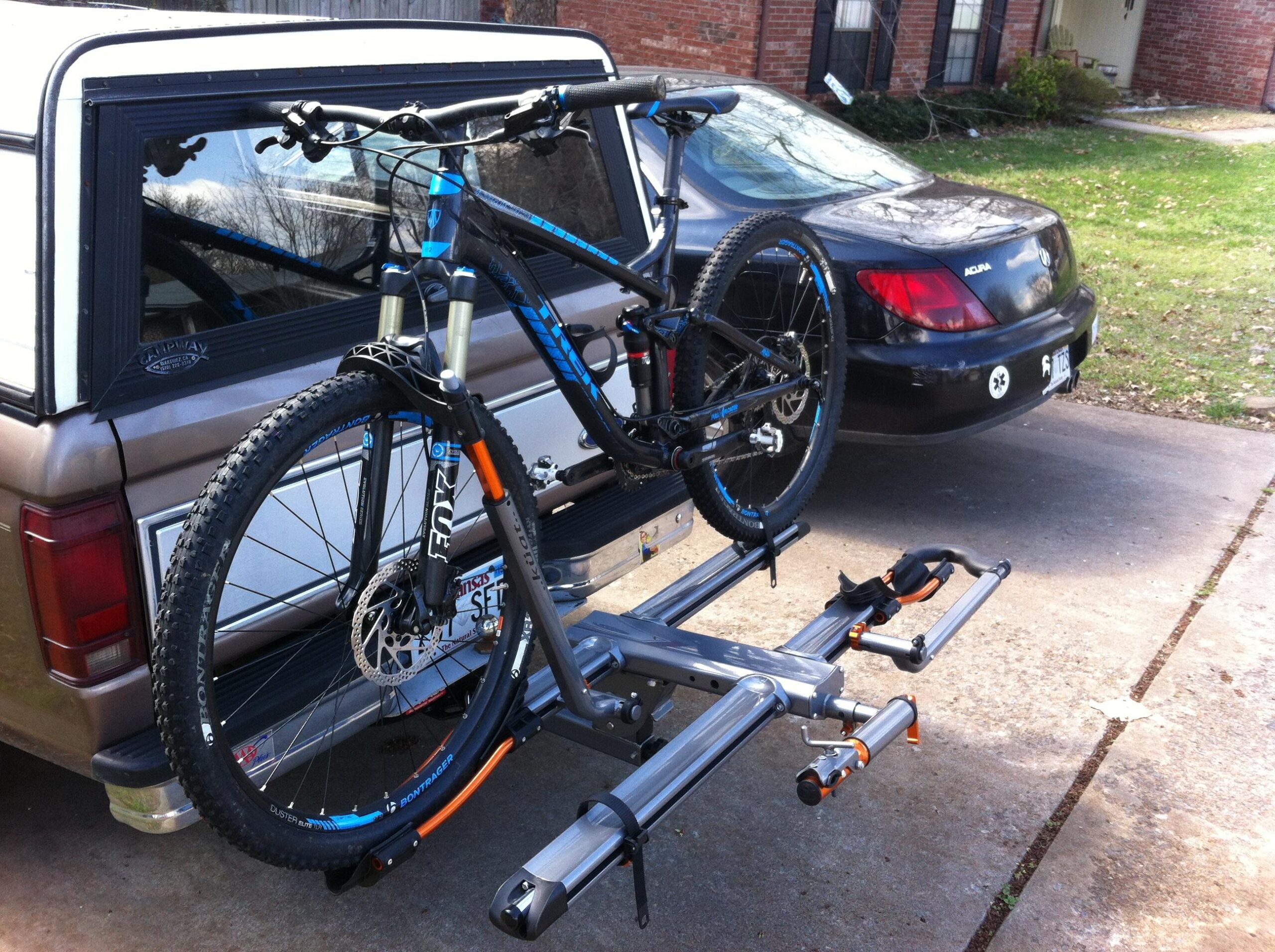 Trek Fuel EX 7: Alt text: A black and blue mountain bike mounted on a rear bike rack attached to a tan pickup truck. The background features a black car and grass area, with a residential setting visible.