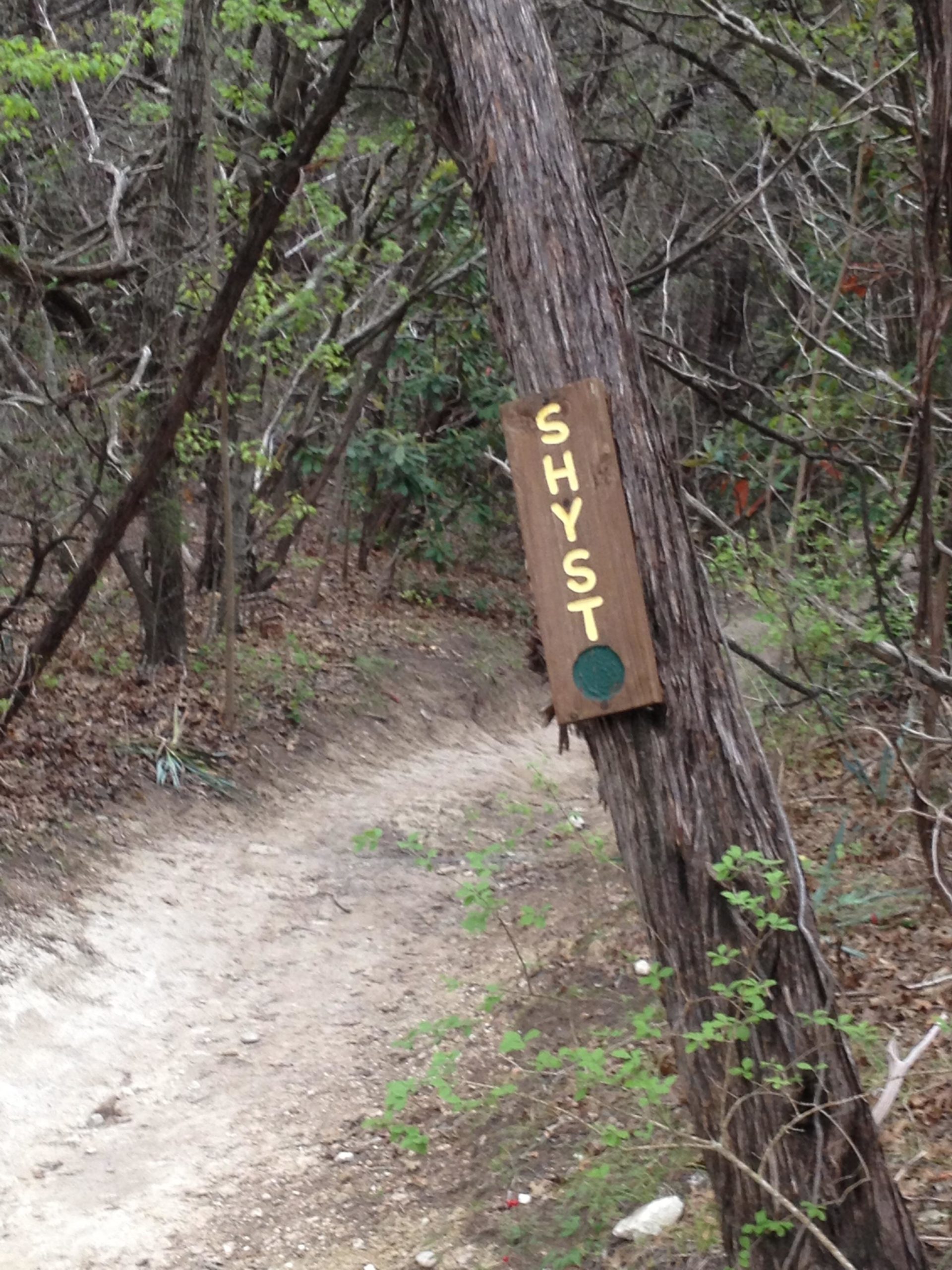 A wooden sign labeled "SHYST" attached to a leaning tree along a dirt path in a wooded area. The background features greenery and sparse underbrush, indicating a natural trail setting. Cameron Park mountain bike trail.