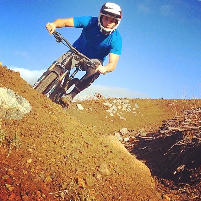 A person in a blue shirt and helmet riding a mountain bike on a dirt trail, leaning into a turn, with a clear blue sky in the background. The terrain is rough with some rocks and dirt. Cold Creek mountain bike trail.