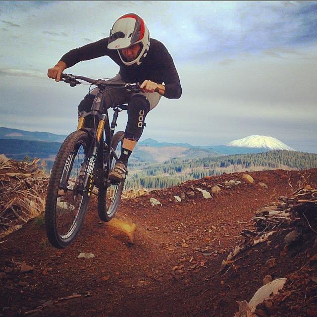 A mountain biker in a helmet performs a jump on a dirt trail, surrounded by forested hills and a snow-capped mountain in the background. The sky is partly cloudy, creating a dynamic outdoor scene. Cold Creek mountain bike trail.