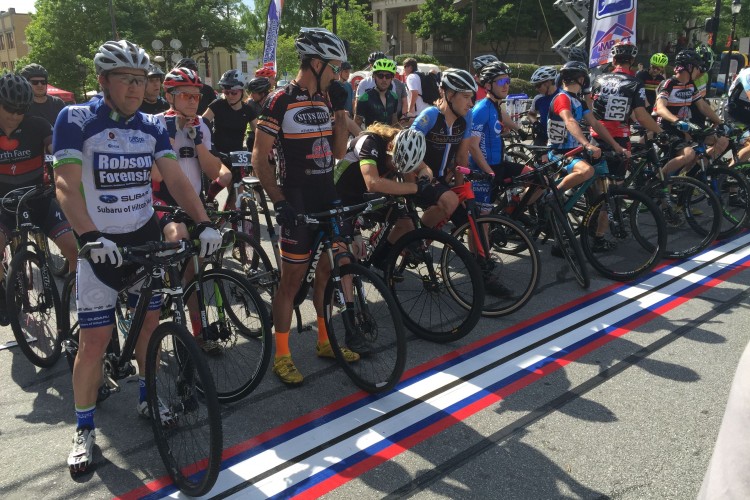 A group of cyclists in various jerseys and helmets are lined up at the starting line of a cycling race. The scene takes place on a sunny day, with a blue sky and trees in the background. Participants are on their bikes, ready for competition, as they prepare to begin the race. The starting line is marked with red, white, and blue stripes on the pavement.