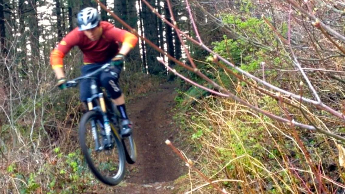 A mountain biker in a red shirt and helmet jumps off a trail surrounded by trees and bushes. The biker is mid-air, showcasing an action shot that captures the excitement of off-road cycling. Cold Creek mountain bike trail.
