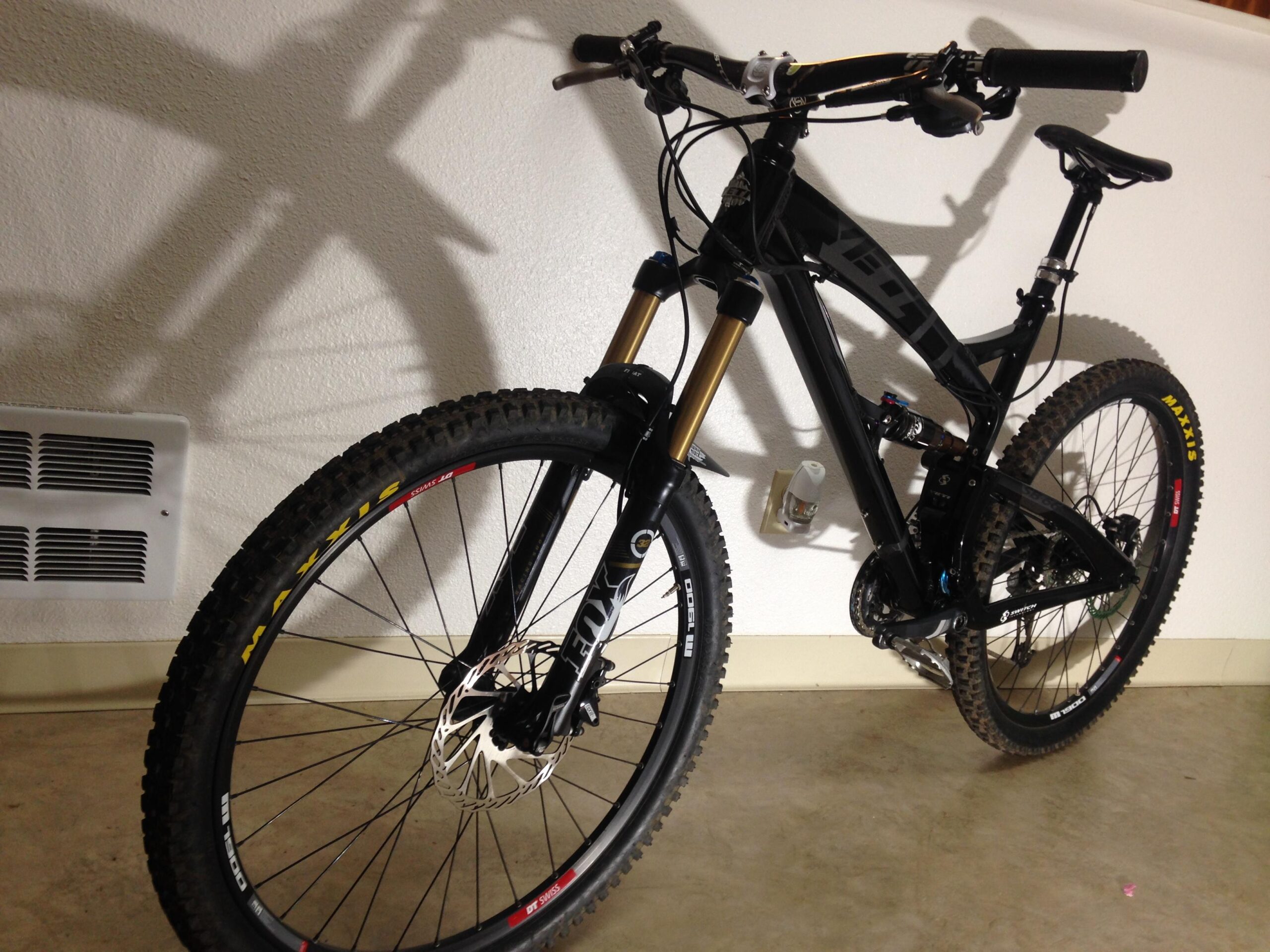 Yeti SB66: A black mountain bike leaning against a wall, featuring thick tires and front suspension. The bike's frame showcases a sleek design, with visible components such as the handlebars, gears, and braking system. Shadows cast on the wall add depth to the image.