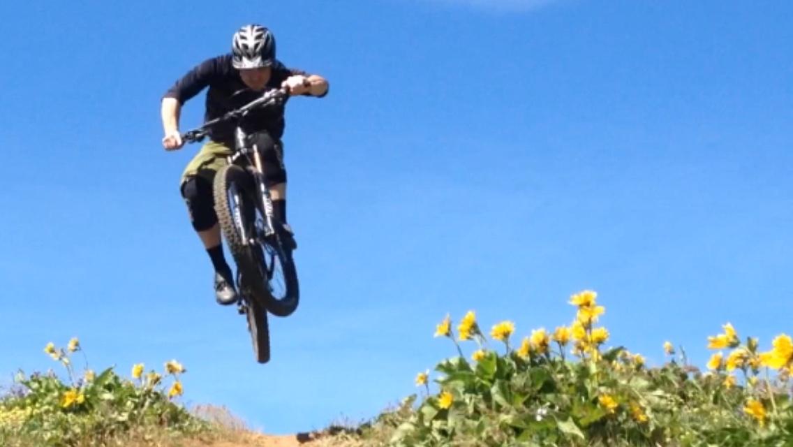 A mountain biker performing a jump over a dirt path, surrounded by yellow wildflowers under a clear blue sky. Syncline mountain bike trail.