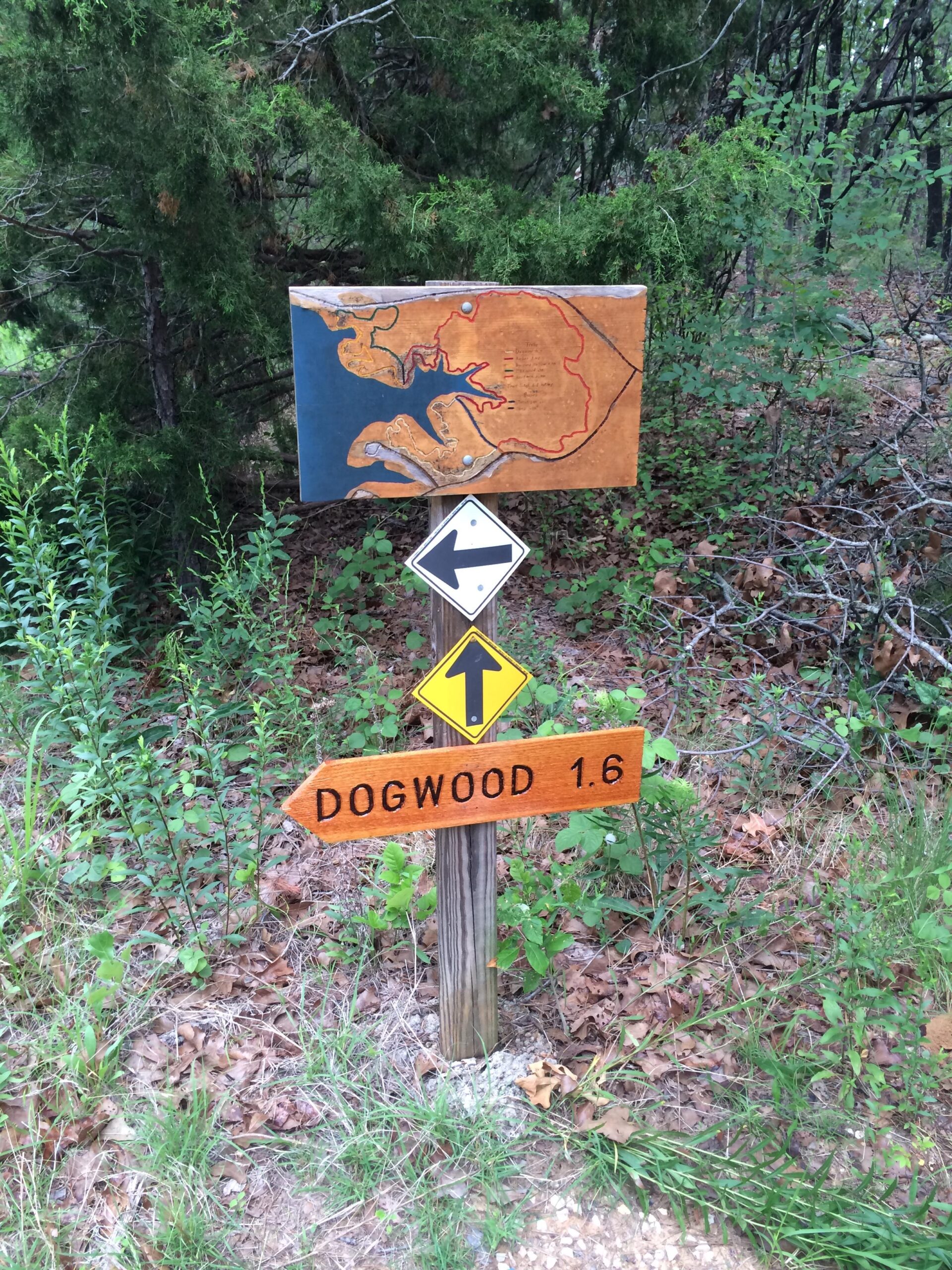 A wooden trail sign showing a map of the area, with a red trail marked. Below the map, there are directional arrows indicating a left turn and straight ahead, with text pointing to "DOGWOOD 1.6," suggesting the distance to the Dogwood trail. Surrounding the sign is dense green foliage and underbrush. Barber Hills Trail at Pat Mayse Lake mountain bike trail.