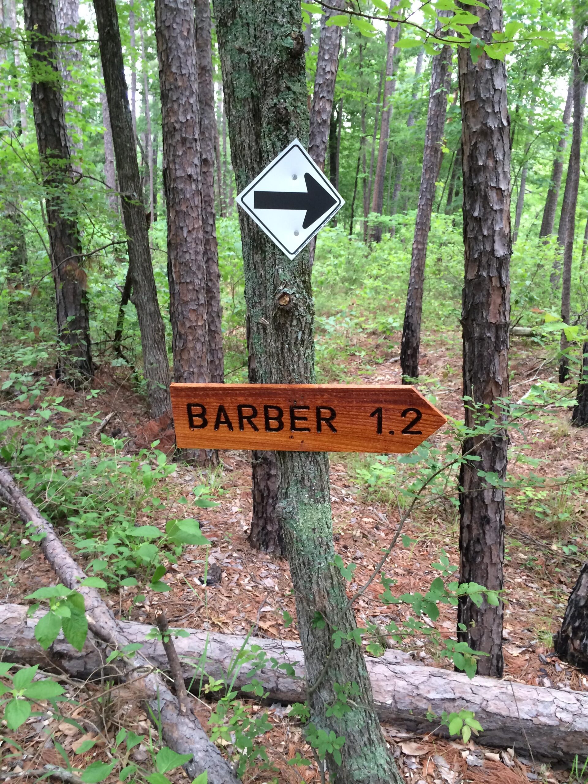 Signpost indicating the direction and distance to "Barber" trail, located in a wooded area with tall pine trees and underbrush. The sign is wooden with black lettering and points to the right, showing a distance of 1.2 miles. Barber Hills Trail at Pat Mayse Lake mountain bike trail.