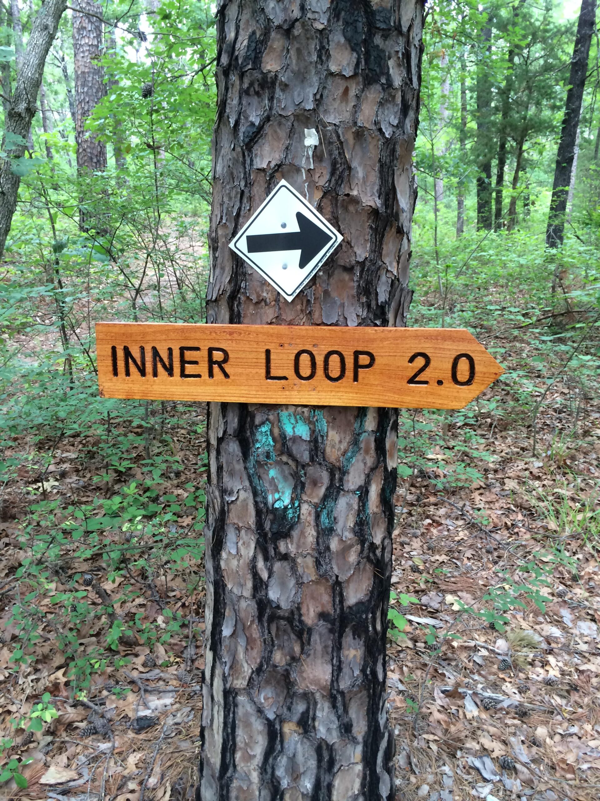 A wooden sign with the text "INNER LOOP 2.0" pointing to the right, attached to a tree in a forested area. A white diamond-shaped sign with a black arrow is positioned above the wooden sign, surrounded by greenery and fallen leaves on the forest floor. Barber Hills Trail at Pat Mayse Lake mountain bike trail.