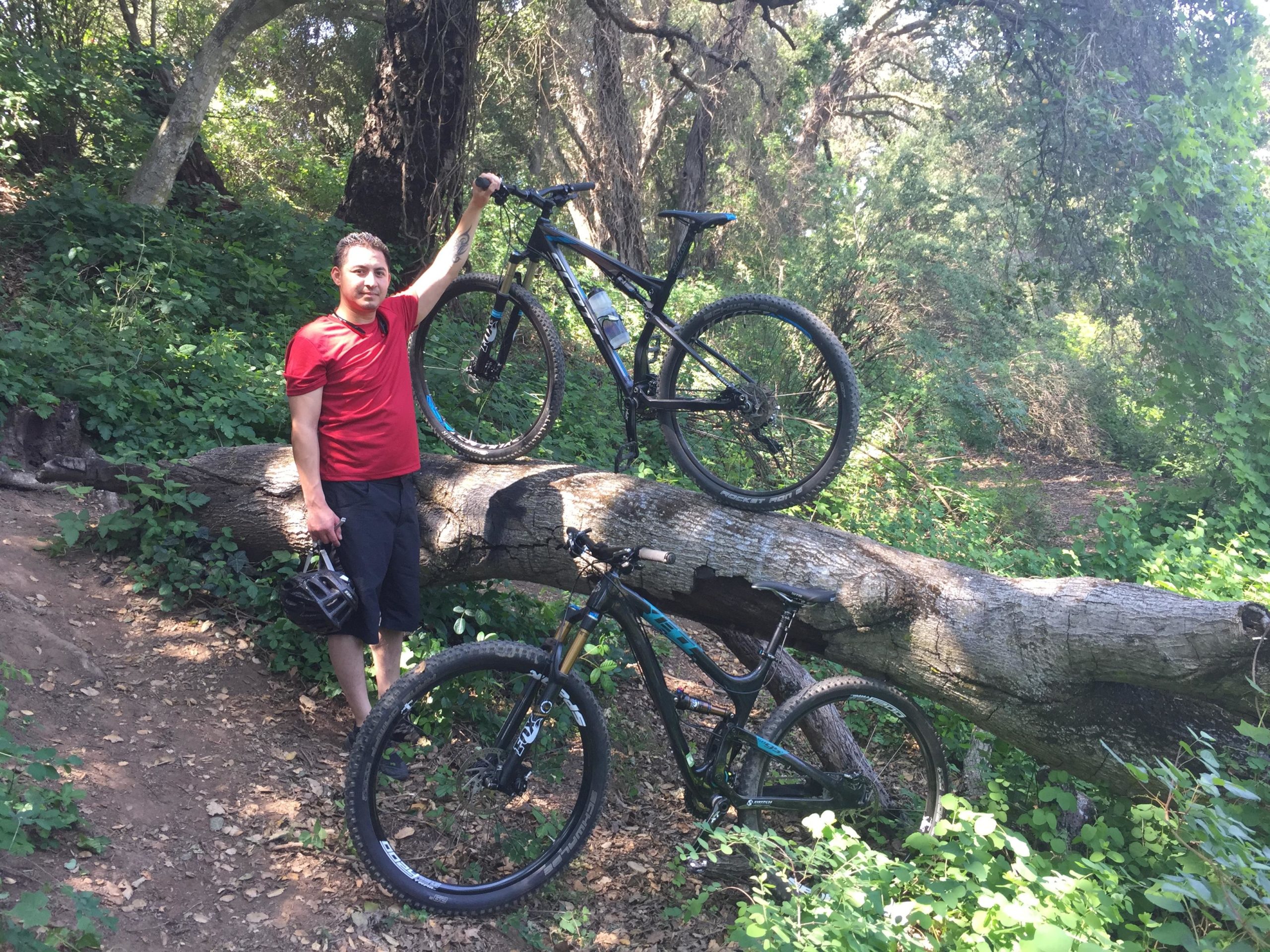 A person in a red shirt stands on a fallen log in a lush, green forest, holding a bike above their head. Another bike is propped against the log beside them. The sun filters through the trees, illuminating the vibrant vegetation around them. Christmas Hill Garlic Ride mountain bike trail.