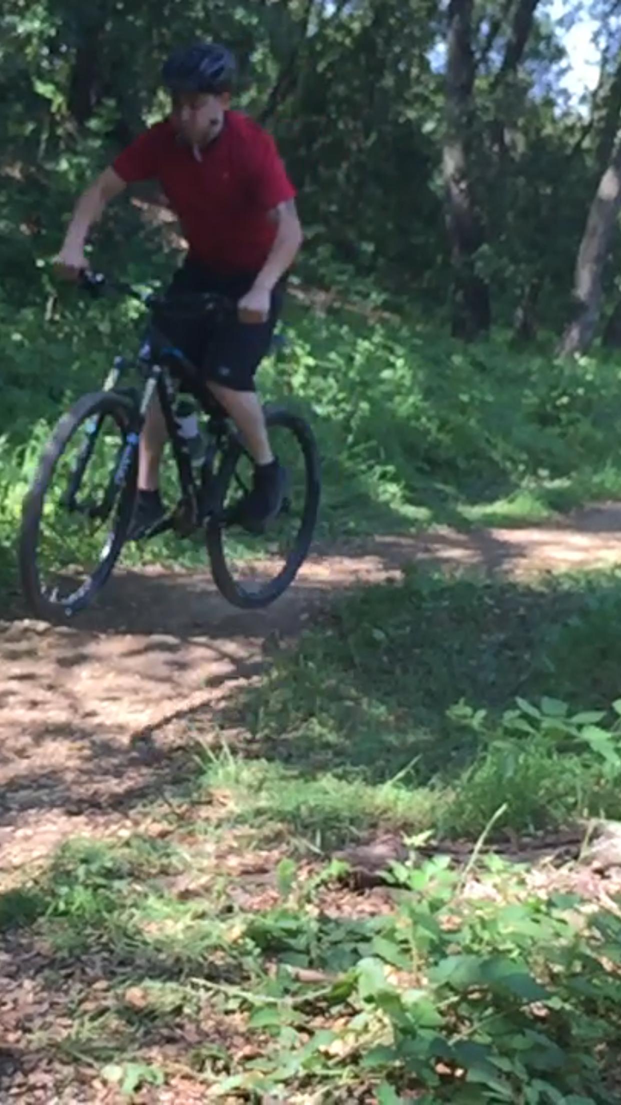 A person wearing a red shirt and a helmet is riding a mountain bike along a dirt trail surrounded by greenery and dense trees. The cyclist is in motion, with one wheel lifted slightly off the ground, suggesting a dynamic ride through a natural landscape. Christmas Hill Garlic Ride mountain bike trail.