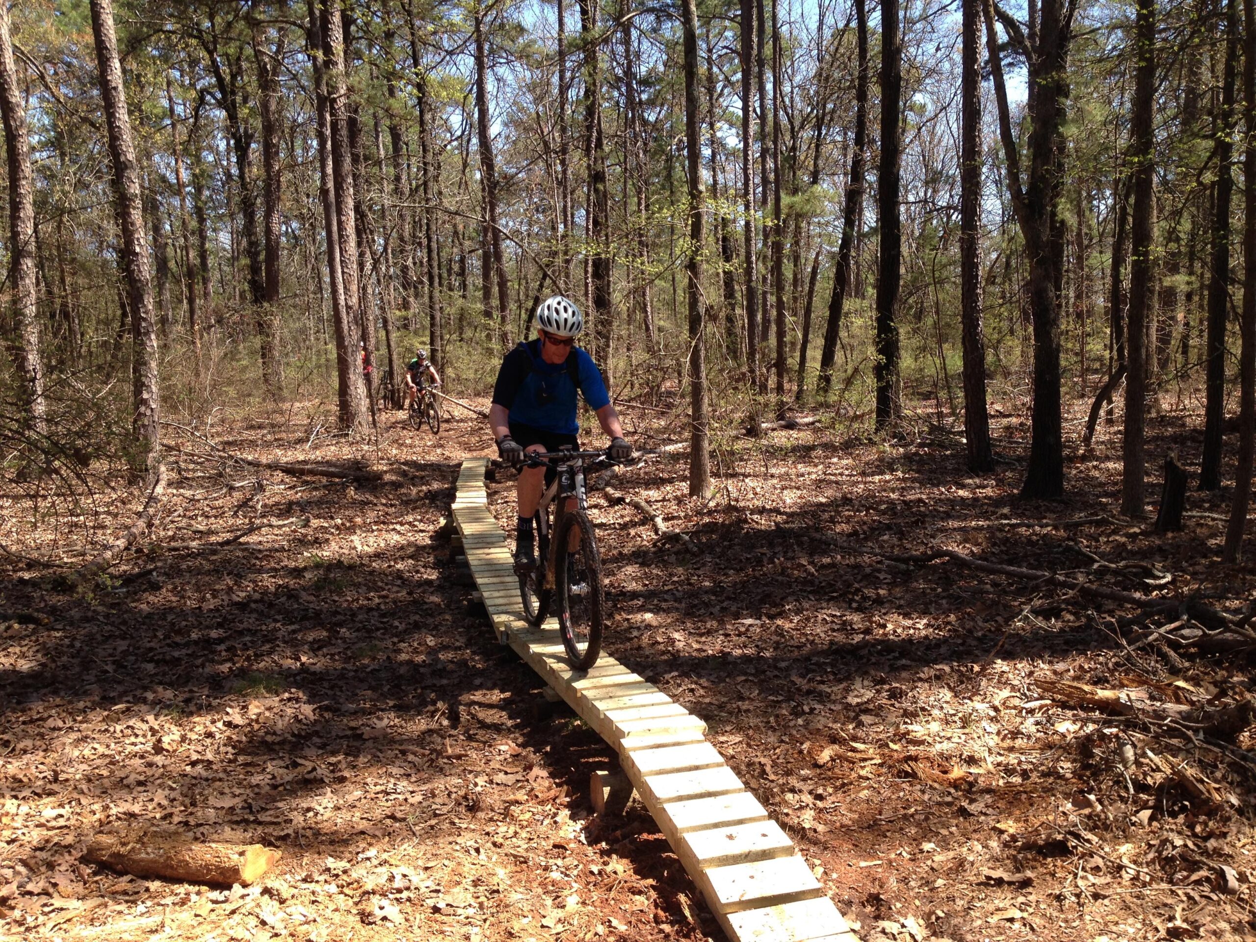 A mountain biker riding on a wooden bridge through a forested trail, surrounded by trees and scattered leaves on the ground, with another cyclist visible in the background. Barber Hills Trail at Pat Mayse Lake mountain bike trail.