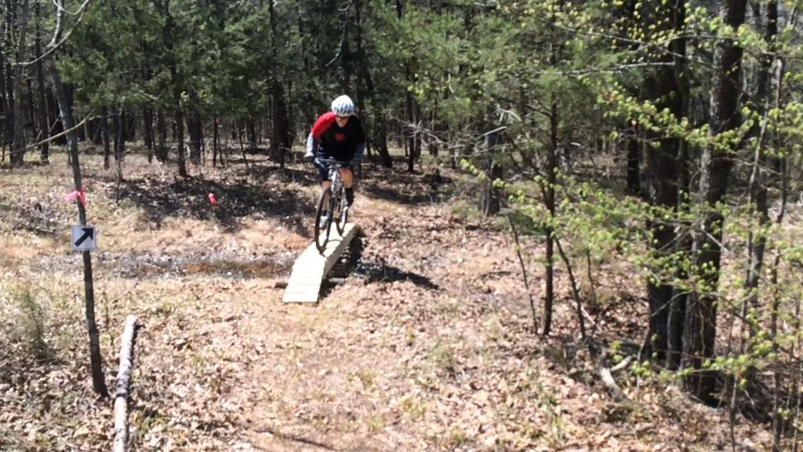 A cyclist in a red and black shirt rides a mountain bike over a wooden ramp on a dirt trail surrounded by trees and underbrush. Pink flags mark the trail direction, and a sign indicating a turn is visible on the left. The scene captures a sunny day in a natural, wooded environment. Barber Hills Trail at Pat Mayse Lake mountain bike trail.
