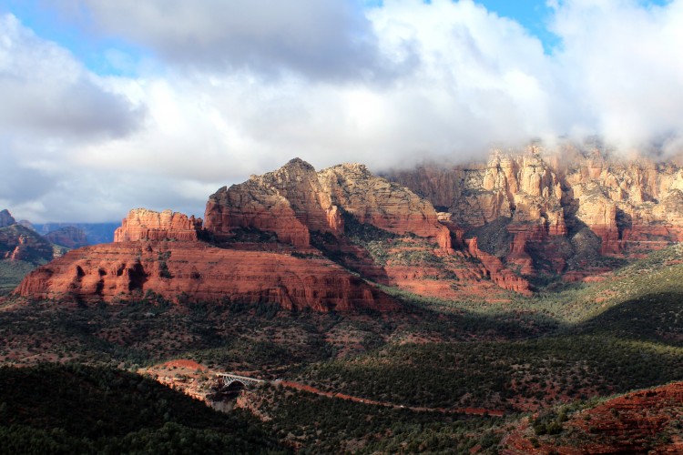 A picturesque view of majestic red rock formations and rugged mountains under a partly cloudy sky. The landscape features lush green vegetation in the foreground, contrasting with the vibrant red hues of the rocks and cliffs in the background, creating a stunning natural scenery.