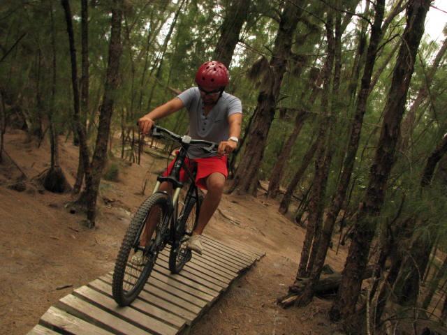 A person riding a mountain bike along a narrow wooden path in a forested area, surrounded by trees. The cyclist is wearing a red helmet and casual clothing, with a focus on balancing while navigating the trail. Oleta River State Park mountain bike trail.