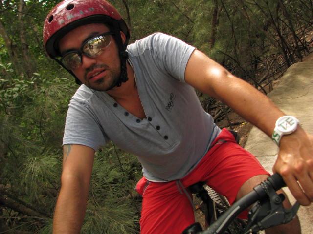 A person wearing a red helmet and sunglasses is riding a bicycle on a wooded path, wearing a light blue shirt and red shorts. The background features greenery and trees, indicating an outdoor cycling activity. Oleta River State Park mountain bike trail.