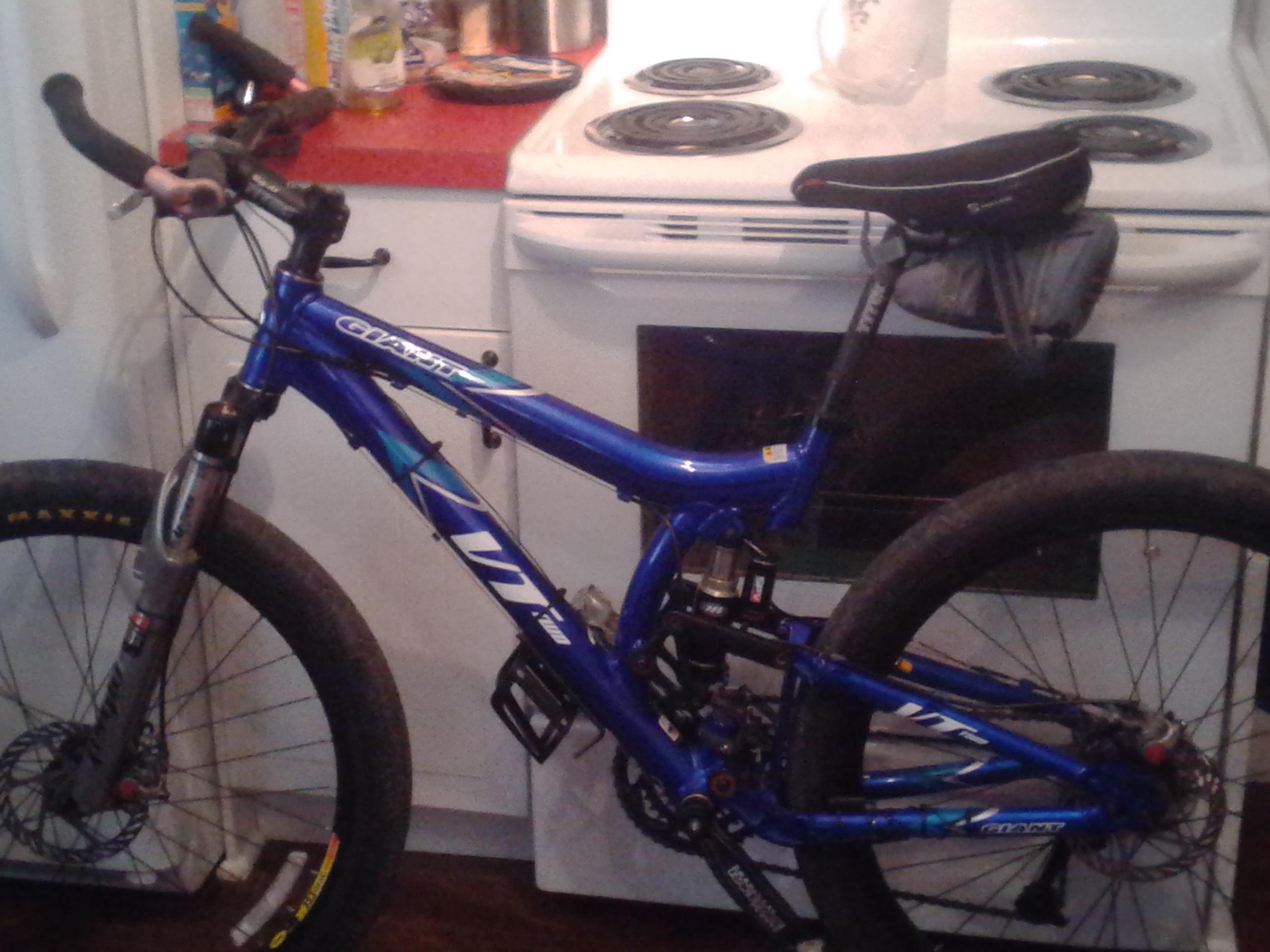 Giant VT2: A blue mountain bike with front suspension and a black saddle bag, positioned in a kitchen setting. The bike rests against a white stove, with a countertop and various kitchen items in the background.