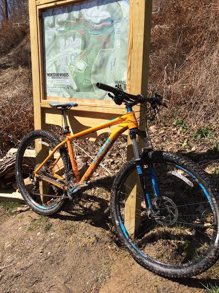 Trek Stache: An orange mountain bike resting against a wooden sign displaying a map of the trails at Montour Woods. The bike is partially muddy, indicating recent use, and the background features natural terrain with dry vegetation.