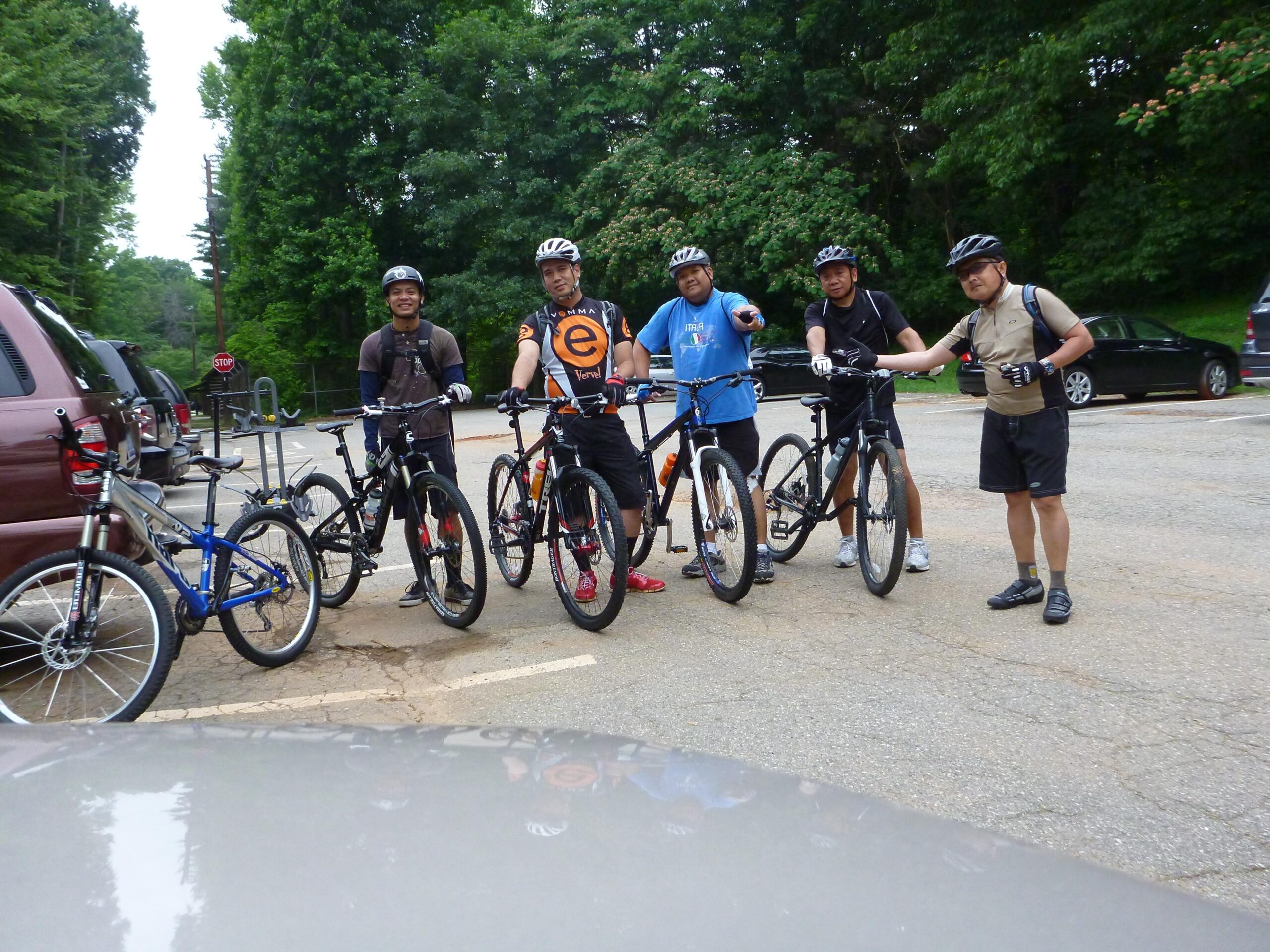 A group of five mountain bikers stands beside their bikes in a parking area, surrounded by trees. They are wearing helmets and cycling gear, with some holding their bikes and others gesturing as they chat. A brown vehicle is partially visible in the foreground. The scene appears to be set in a recreational area, suggesting an outing or ride together. Country Park mountain bike trail.