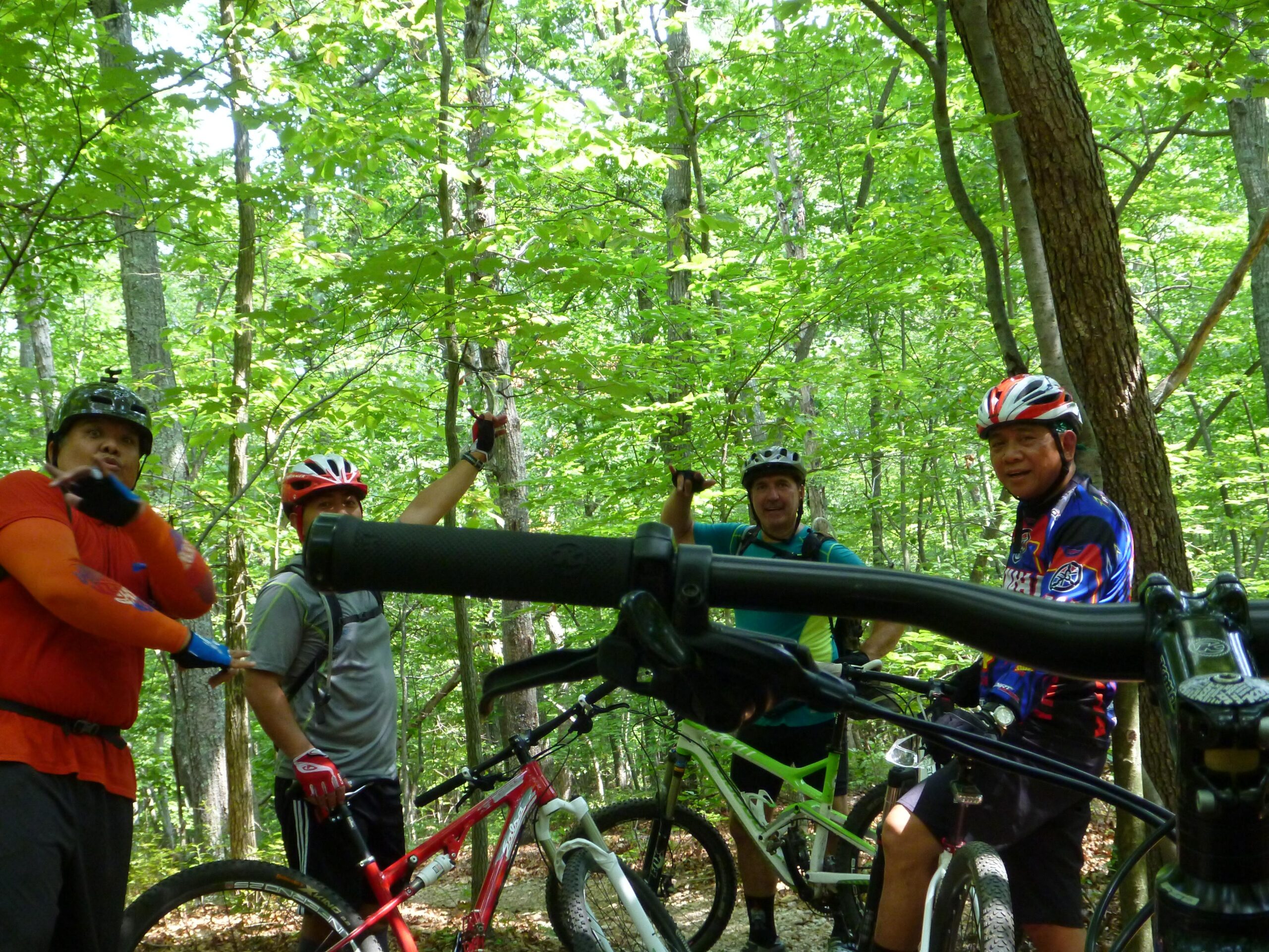 A group of four mountain bikers gathered in a lush green forest, each wearing helmets and cycling gear. They are standing near their bikes, with two riders gesturing and smiling, while another holds a camera. The scene captures a moment of camaraderie and enjoyment in nature. Country Park mountain bike trail.