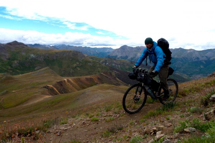 A cyclist navigating a rugged mountain landscape, with rolling hills and distant peaks under a partly cloudy sky. The rider is wearing a blue jacket and a beanie, carrying a backpack and bike gear as they traverse a dirt path amidst wildflowers.