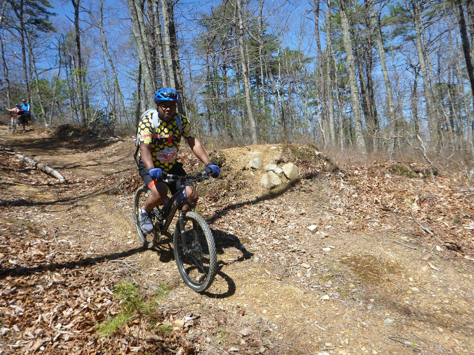 A person riding a mountain bike down a dirt trail in a wooded area, surrounded by trees and scattered leaves on the ground. The cyclist is wearing a blue helmet and a colorful patterned jersey. Another cyclist can be seen in the background, also navigating the trail. The sky is clear and blue, indicating a sunny day. Carvin's Cove Trail system mountain bike trail.