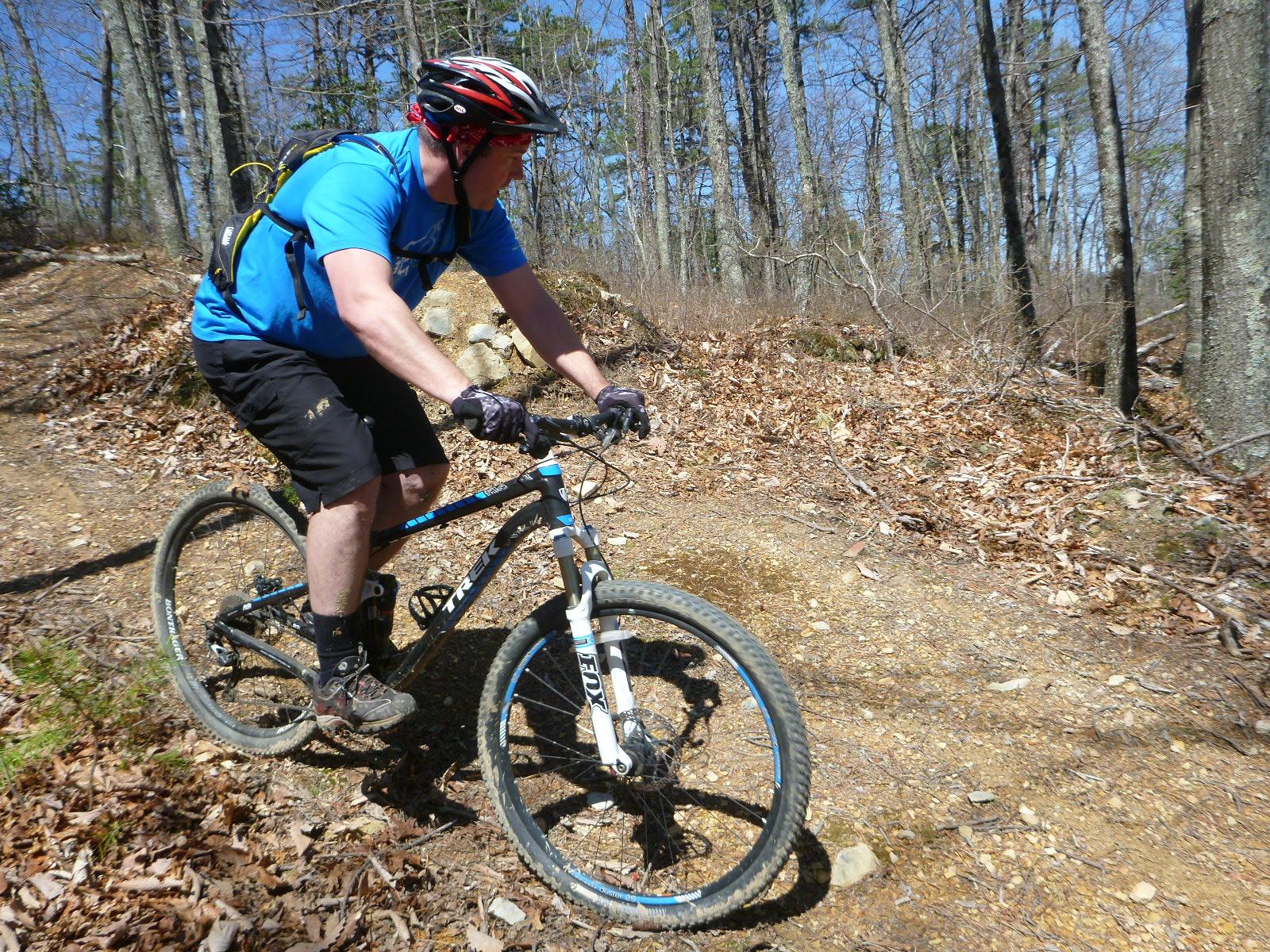 A person riding a mountain bike on a wooded trail during sunny weather. The rider is wearing a blue shirt, black shorts, and a helmet with a backpack, navigating a path covered in leaves and dirt. Trees line the trail in the background. Carvin's Cove Trail system mountain bike trail.