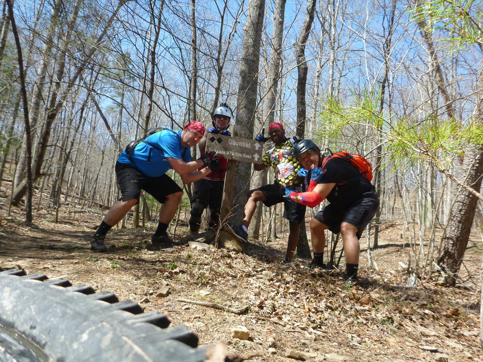 Four friends posing enthusiastically beside a trail sign in a wooded area. They are wearing biking gear, including helmets and colorful shirts, with smiles and playful poses. The background features trees and a clear blue sky, suggesting a sunny day outdoors. Carvin's Cove Trail system mountain bike trail.