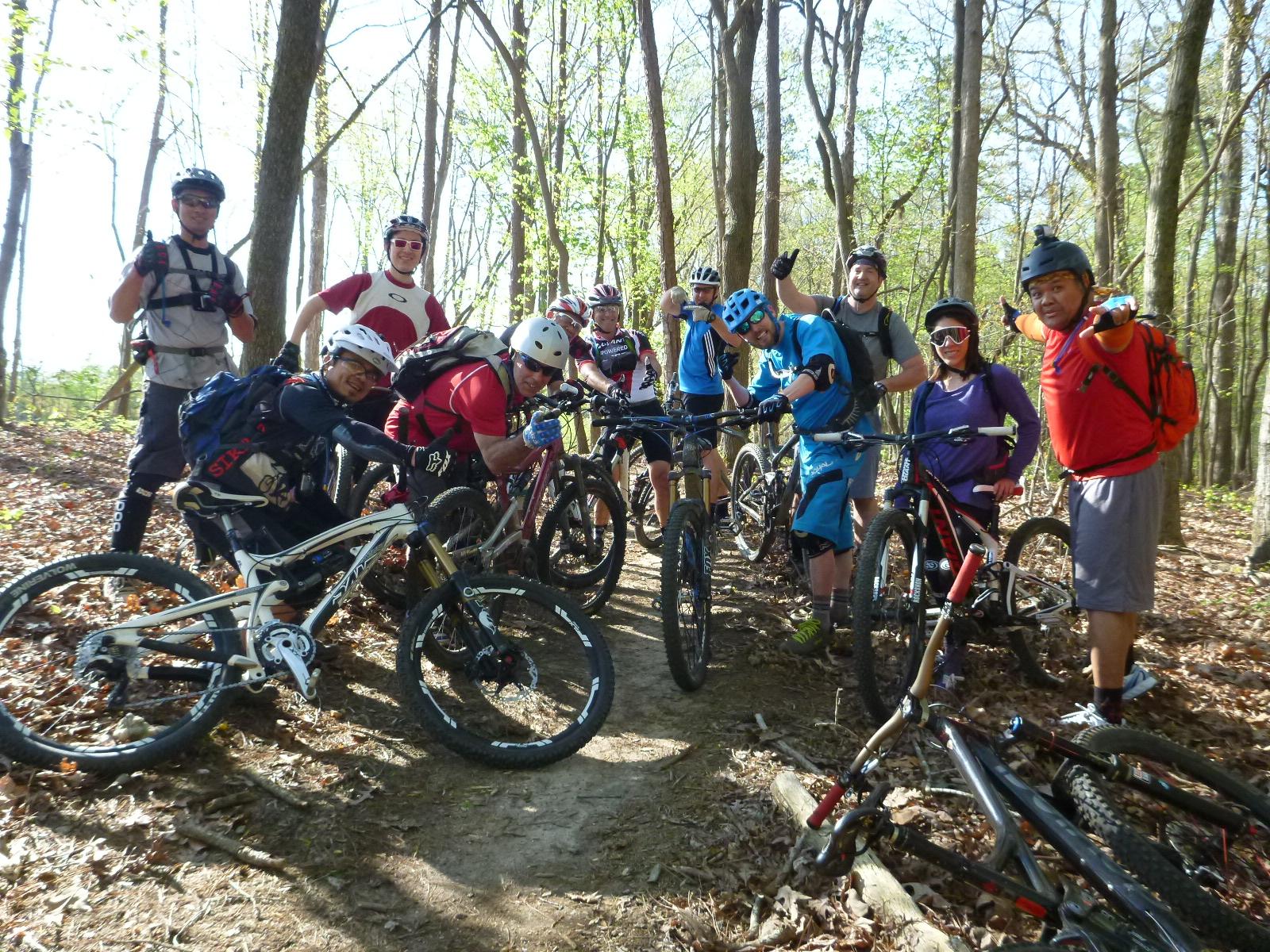 A group of mountain bikers posing for a photo on a forest trail, surrounded by trees and fallen leaves. They are wearing helmets and colorful biking gear, with their bicycles parked nearby. The atmosphere is cheerful and energetic, capturing a moment of camaraderie among the riders. San-lee Park mountain bike trail.