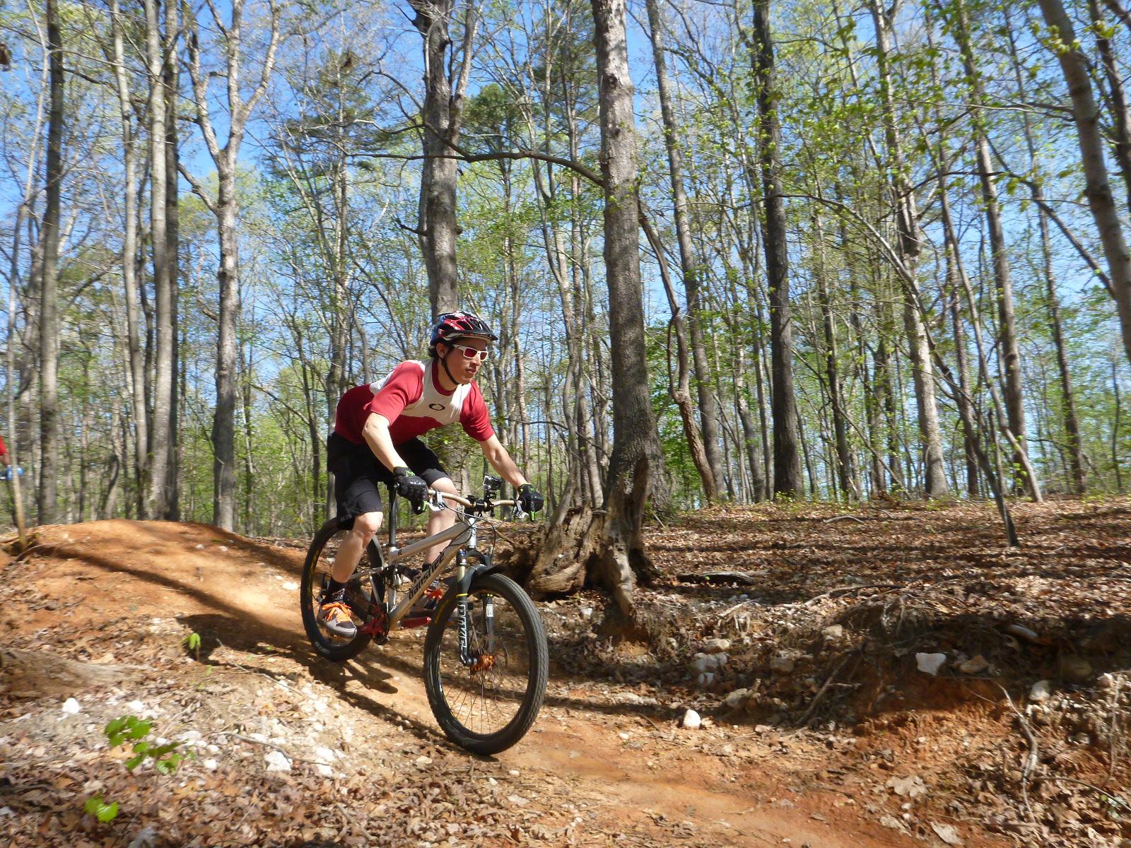 A mountain biker in a red and white jersey rides along a dirt trail in a forested area, surrounded by trees and greenery on a sunny day. The trail features a slight elevation, with the cyclist leaning forward as they navigate the path. San-lee Park mountain bike trail.