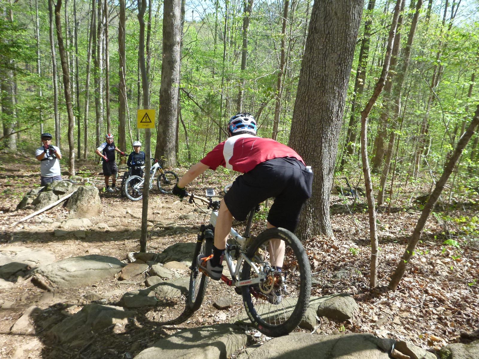 A mountain biker in a red shirt navigates a rocky trail in a wooded area, while other bikers and spectators watch nearby. A warning sign is visible in the background, indicating the trail's difficulty. The scene is set in a lush green environment with trees and underbrush. San-lee Park mountain bike trail.