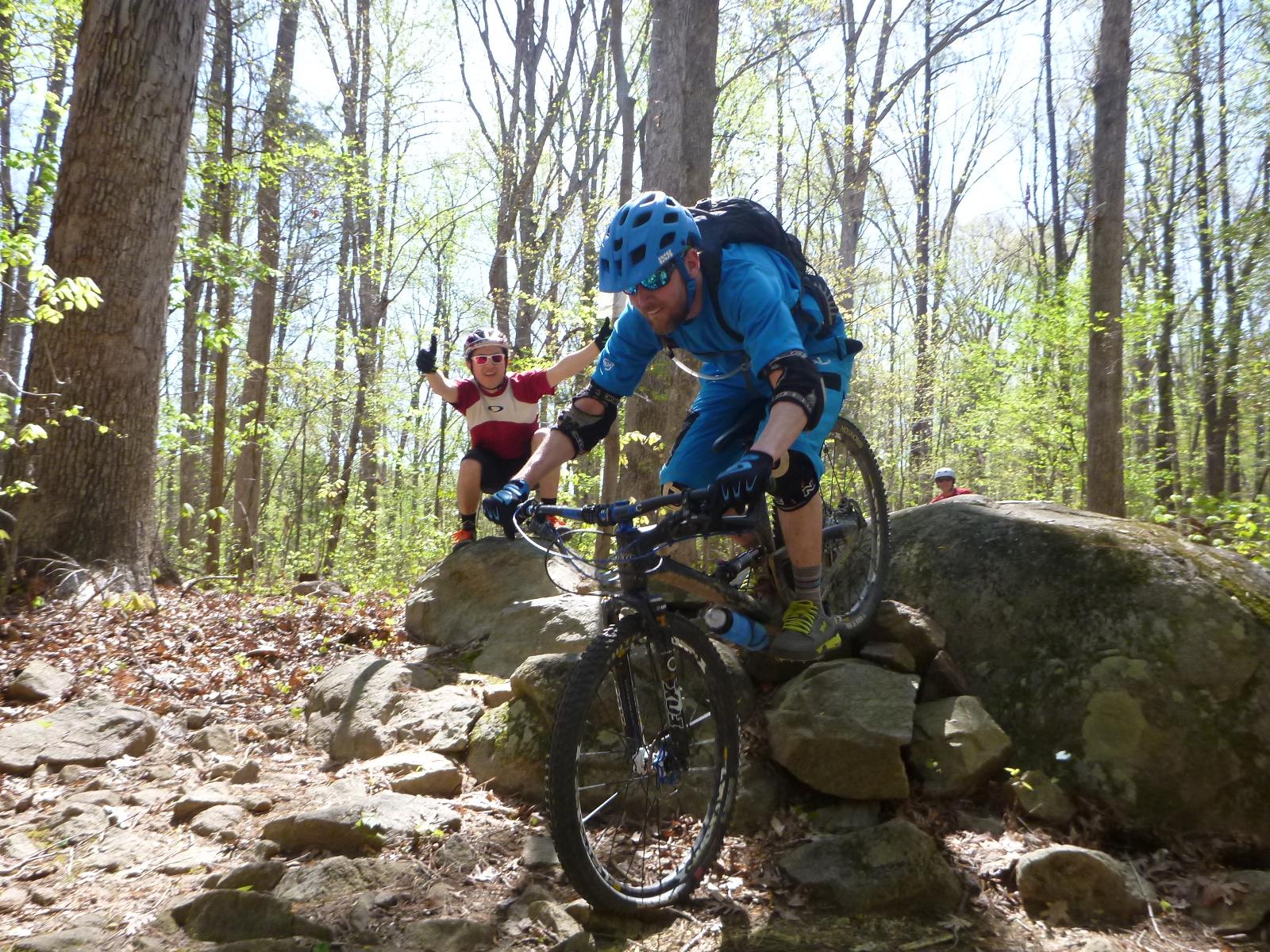 A mountain biker in a blue jersey and helmet navigates over a rocky terrain in a forest, while a child in a red and white shirt enthusiastically gives a thumbs-up from a nearby rock. The scene captures a sunny day with lush green trees in the background. The biker appears focused on maintaining balance as he rides over the challenging path. San-lee Park mountain bike trail.