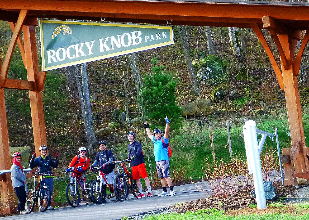 A group of six mountain bikers stands in front of the "Rocky Knob Park" sign, smiling and celebrating their outdoor adventure. They are dressed in casual biking attire and helmets, with several bicycles lined up beside them against a backdrop of trees and rocky terrain. The atmosphere is lively, indicating enjoyment and camaraderie among the riders. Rocky Knob Park mountain bike trail.