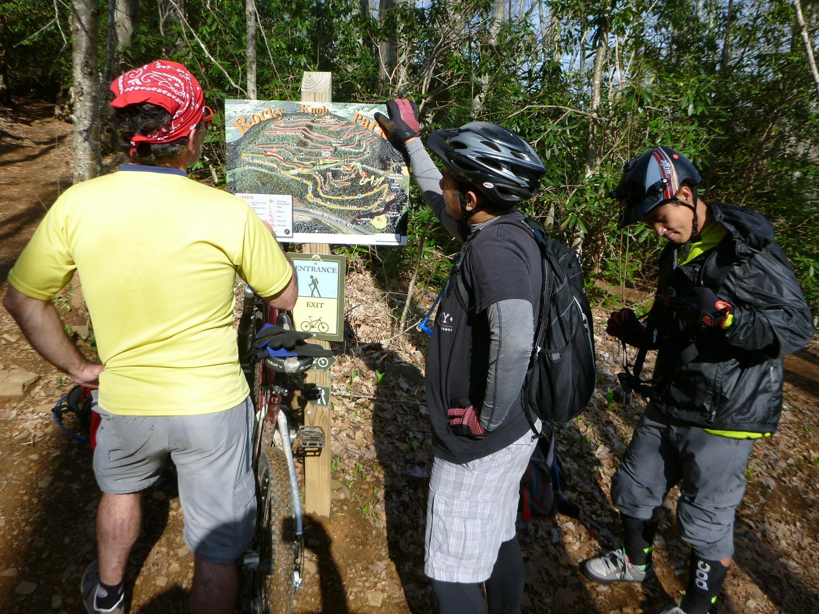 Three mountain bikers stand near a trail map in a wooded area. One biker is pointing at the map, while another is looking at it from behind, and the third is checking equipment. They are dressed in biking gear and helmets, and their bikes are resting nearby. The scene captures a moment of exploration and planning before hitting the trails. Rocky Knob Park mountain bike trail.