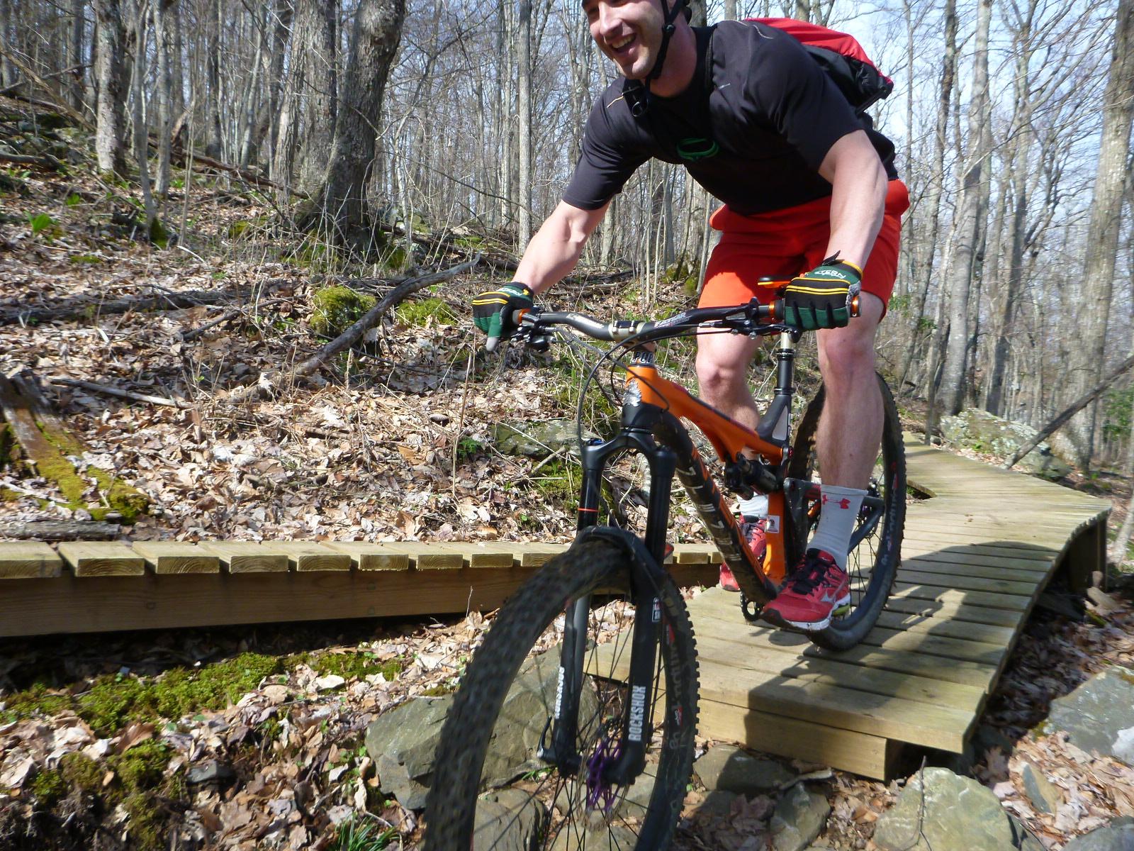 A mountain biker riding on a wooden bridge through a forested area, with trees in the background and fallen leaves on the ground. The rider is wearing a black shirt, red shorts, and gloves, smiling as he navigates the trail. Rocky Knob Park mountain bike trail.
