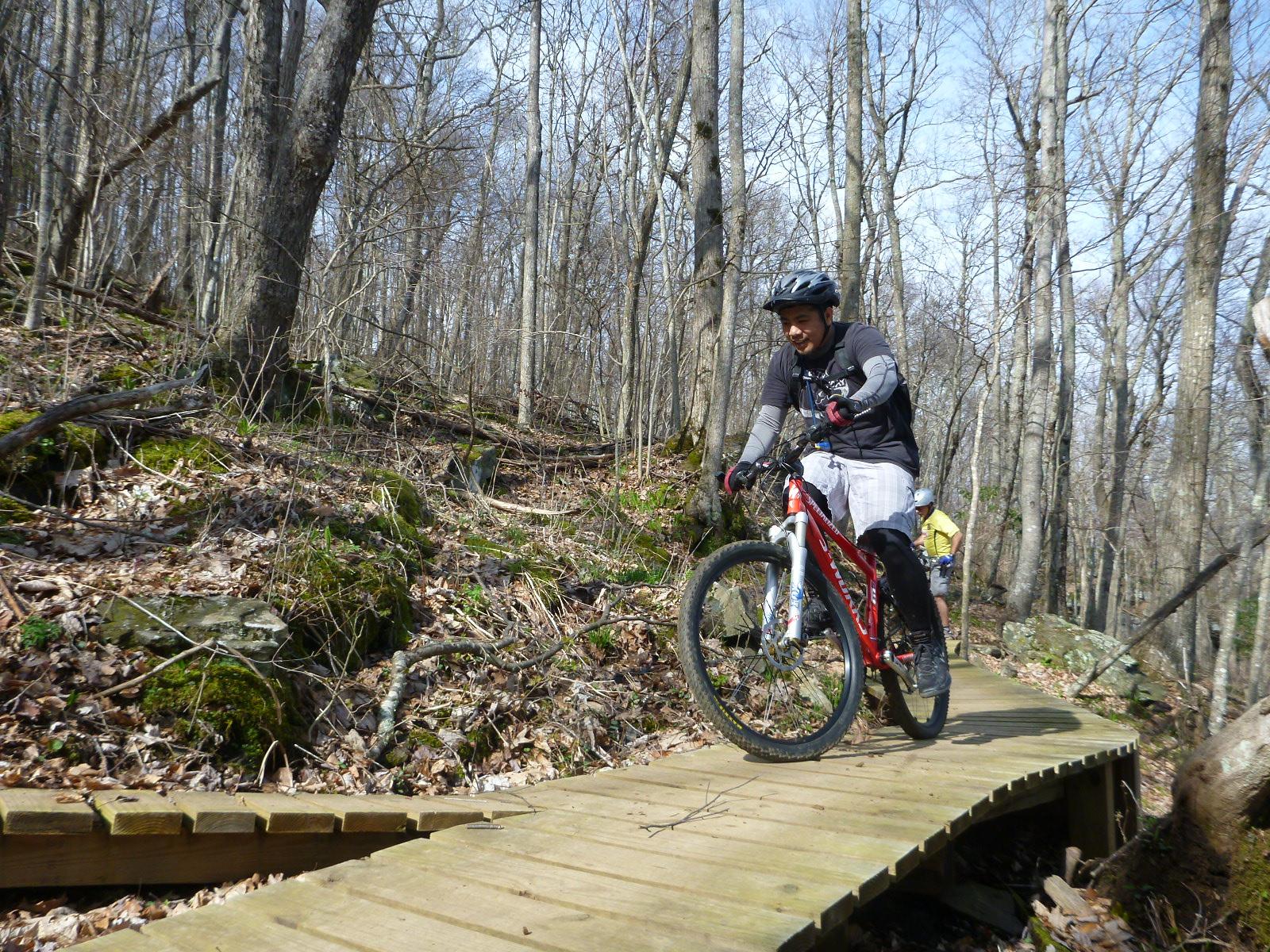 A mountain biker rides along a wooden trail in a forested area, surrounded by bare trees and fallen leaves. The cyclist is wearing a helmet and protective gear, focusing on navigating the path, while another cyclist is visible in the background. The scene captures the essence of outdoor adventure and nature. Rocky Knob Park mountain bike trail.