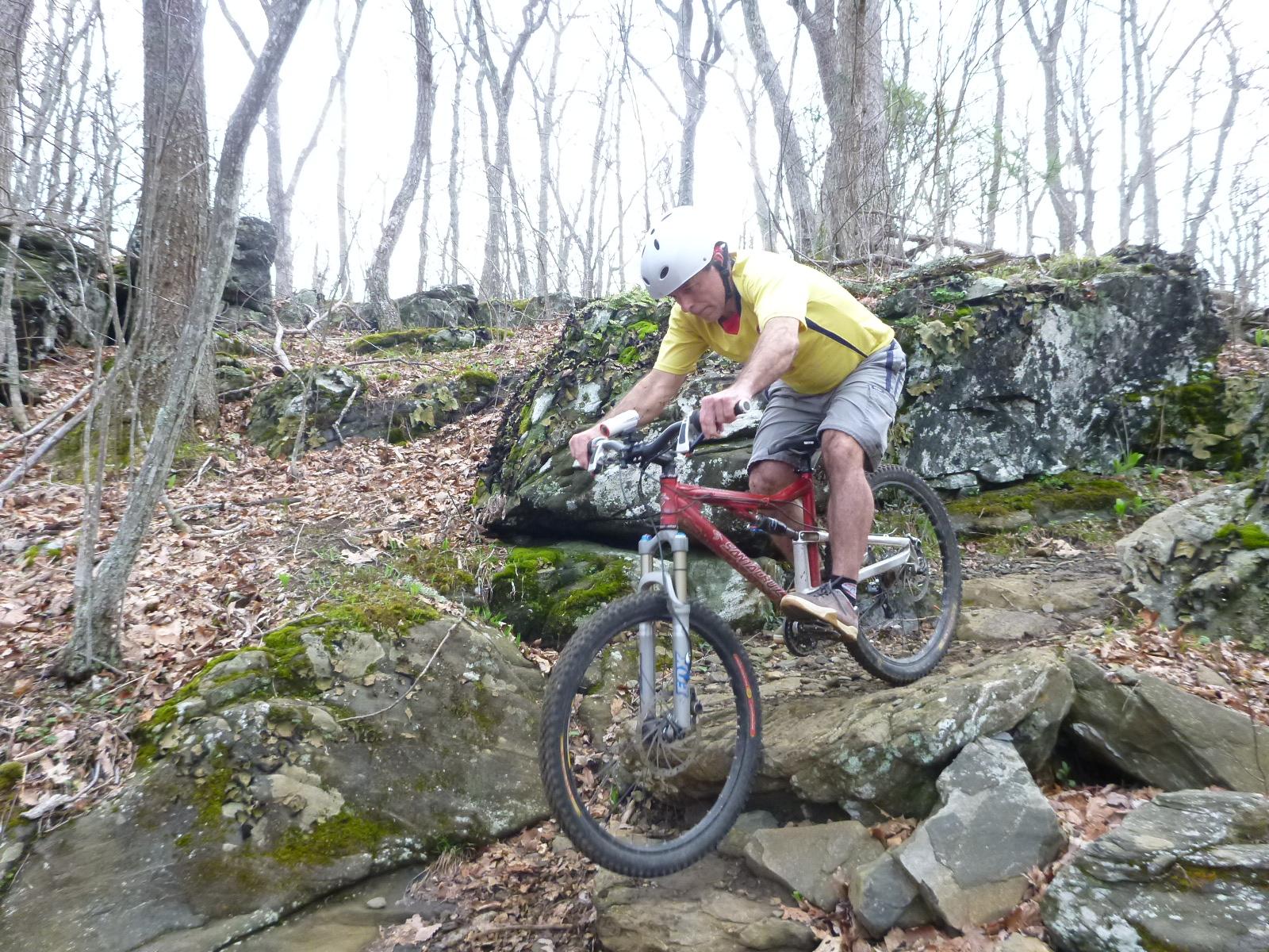 A mountain biker in a yellow shirt and helmet navigates rocky terrain in a wooded area, showcasing an intense moment of riding over rocks and uneven ground. The surrounding environment features bare trees and scattered leaves, indicating a natural trail. Rocky Knob Park mountain bike trail.