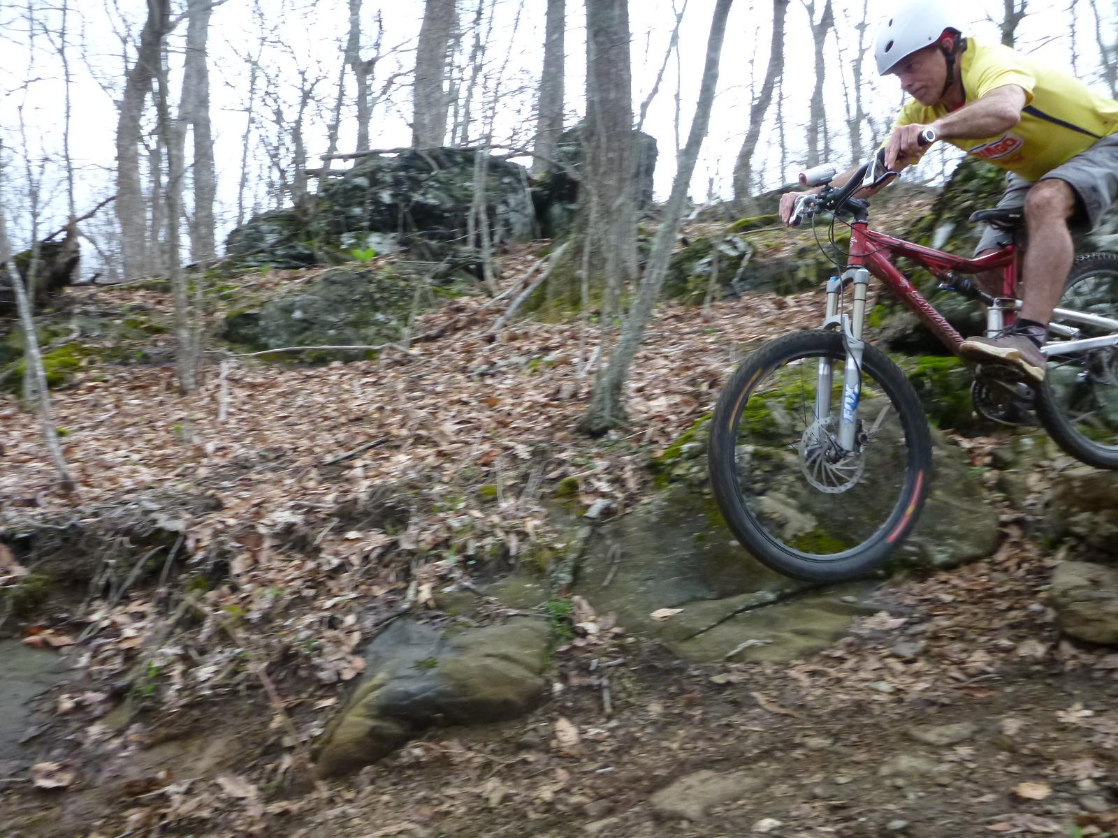 A mountain biker in a yellow shirt and helmet is jumping off a rocky terrain in a wooded area, with trees in the background and fallen leaves on the ground. The biker is positioned mid-air, demonstrating a dynamic riding stance. Rocky Knob Park mountain bike trail.