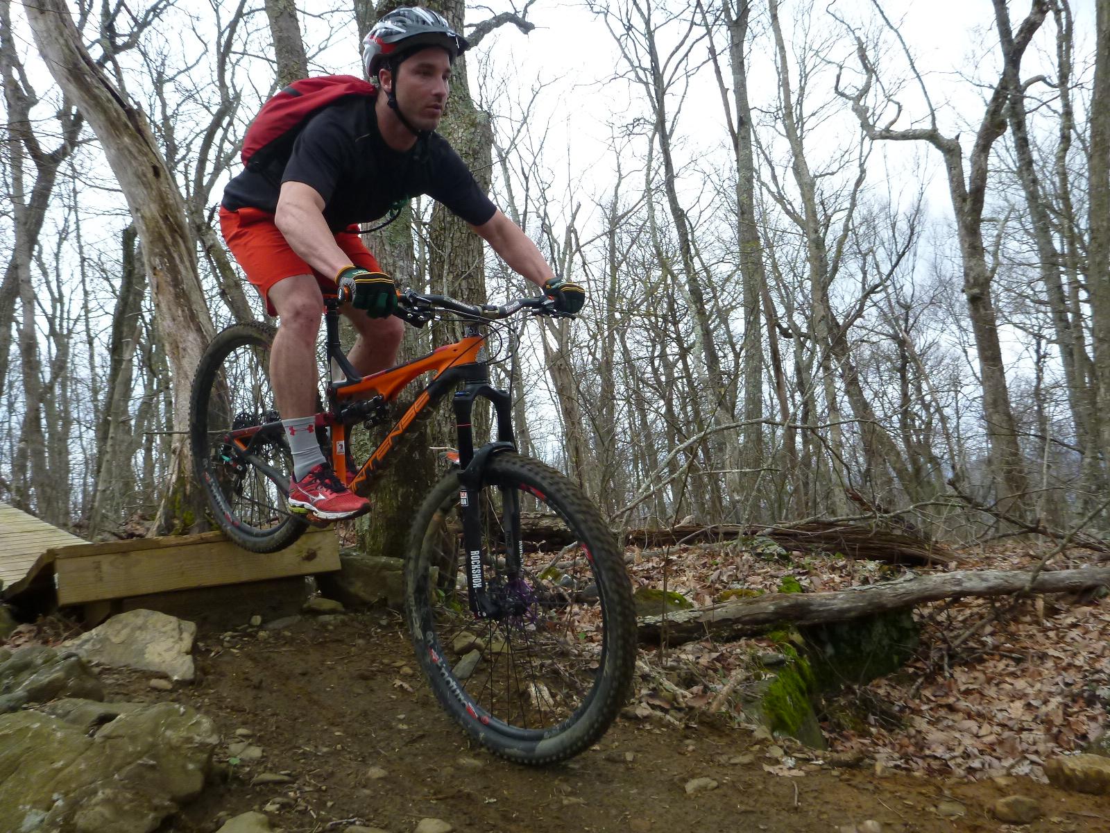 A mountain biker in mid-air jumps over a small wooden ramp on a dirt trail surrounded by trees in a forested area. The rider is wearing a helmet, a black t-shirt, and bright orange shorts, showcasing an action-packed moment of mountain biking. Rocky Knob Park mountain bike trail.