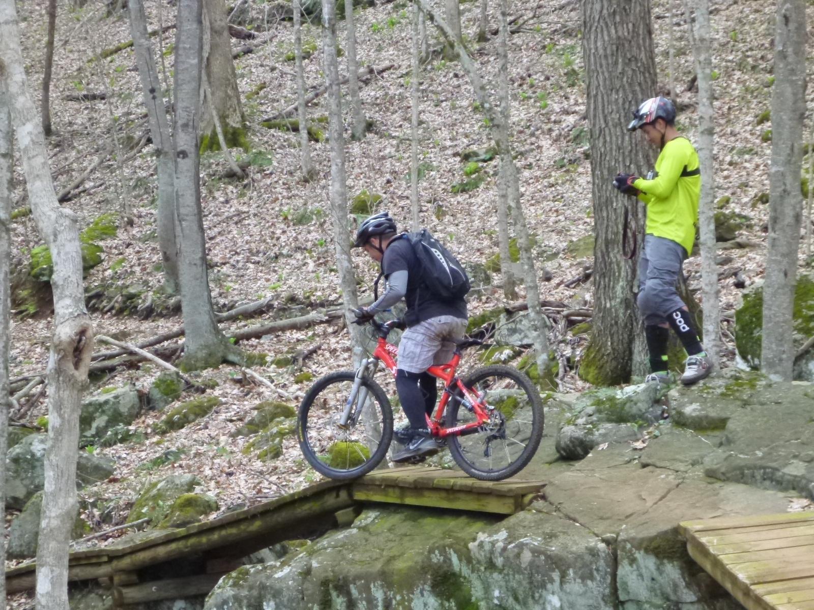 A mountain biker navigating a narrow wooden bridge over rocky terrain in a forested area, while another person in bright yellow clothing stands nearby, observing and recording the ride. Rocky Knob Park mountain bike trail.