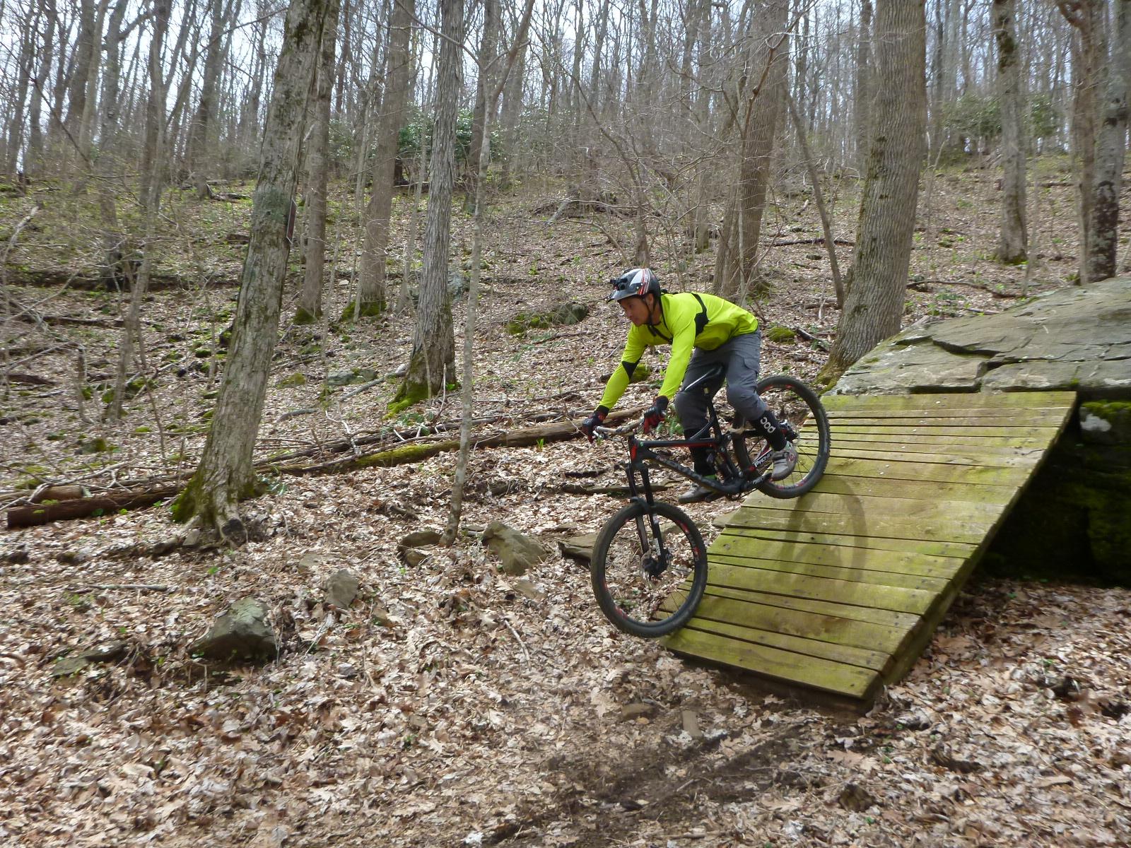 A mountain biker in a bright green jacket descends a wooden ramp in a forested area, surrounded by bare trees and fallen leaves. Rocky Knob Park mountain bike trail.