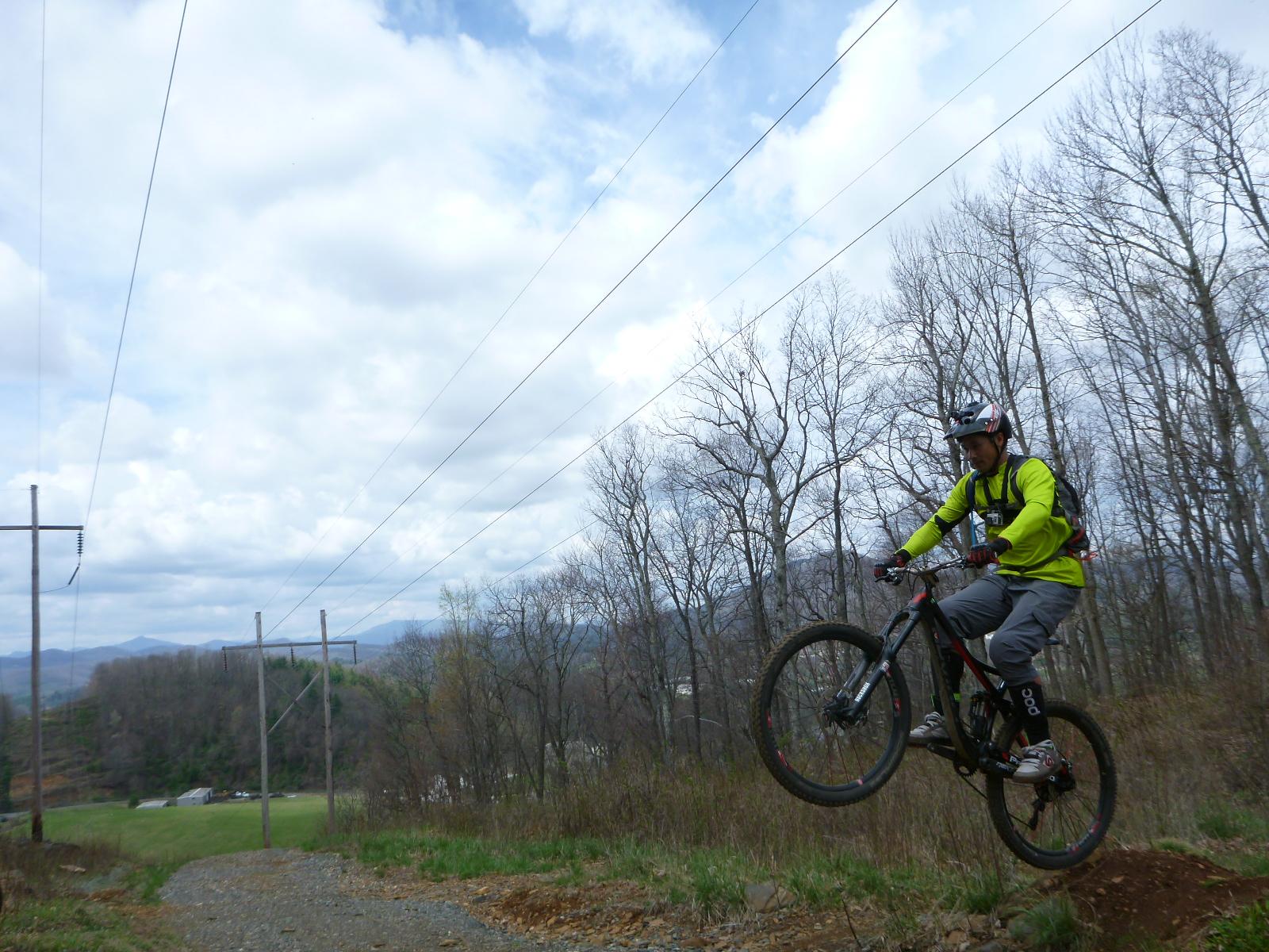 A cyclist in a bright green jacket performs a jump on a mountain bike along a gravel trail surrounded by bare trees and power lines, with rolling hills in the background under a cloudy sky. Rocky Knob Park mountain bike trail.