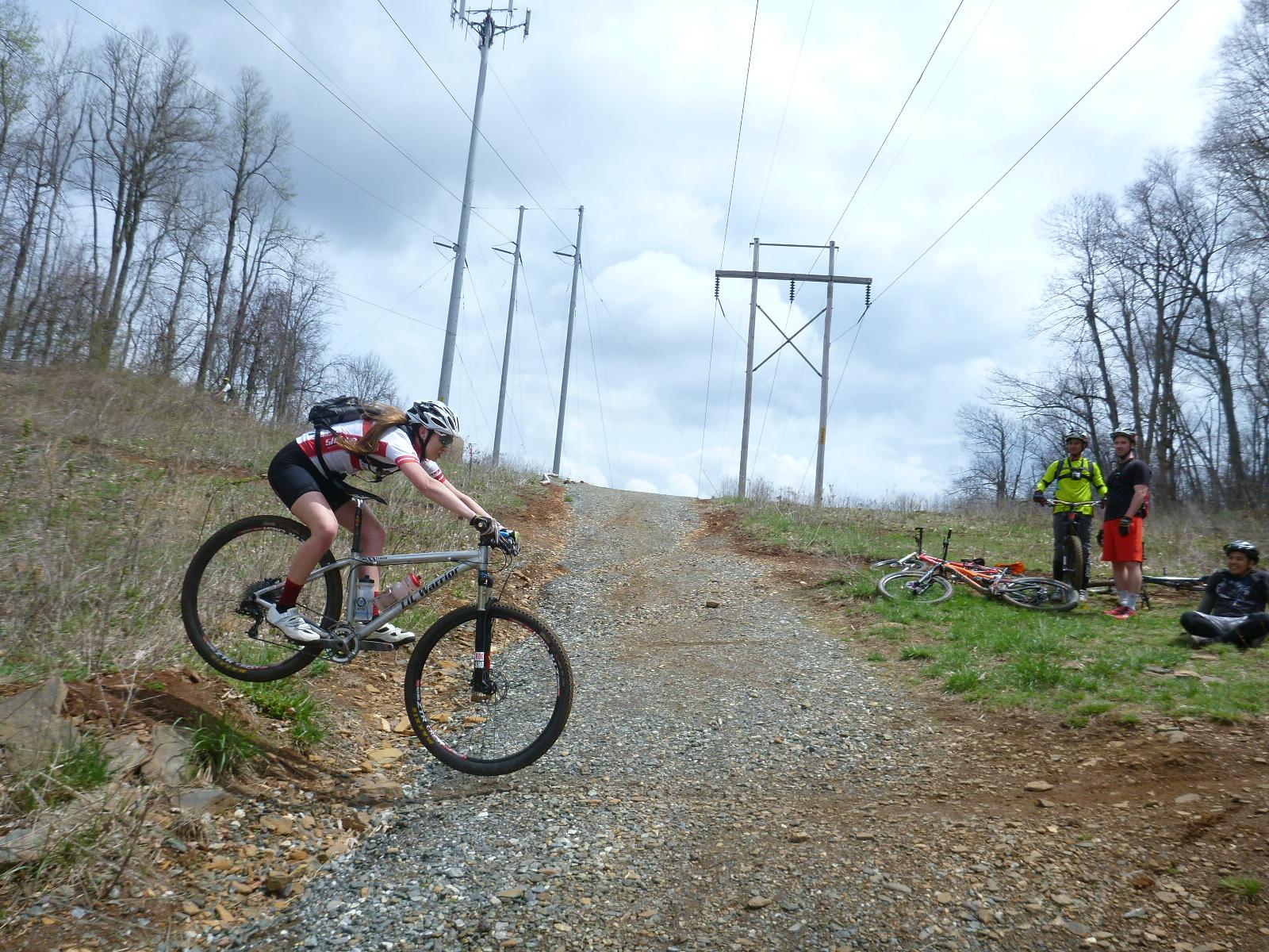 A mountain biker jumps over a small incline on a gravel trail, while three other cyclists observe nearby. The scene is set in a wooded area with power lines in the background and a partly cloudy sky. The cyclist is wearing a helmet and riding attire, showing an active and adventurous atmosphere. Rocky Knob Park mountain bike trail.