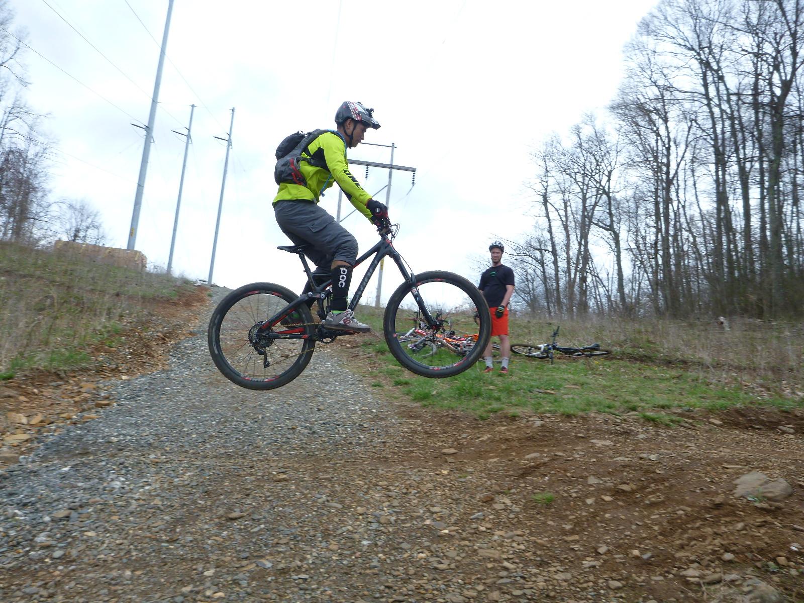 A mountain biker wearing a bright yellow jacket performs a jump on a gravel path, with power lines and trees in the background. Another cyclist in a black shirt and orange shorts watches nearby, while several bicycles are laid on the ground. Rocky Knob Park mountain bike trail.