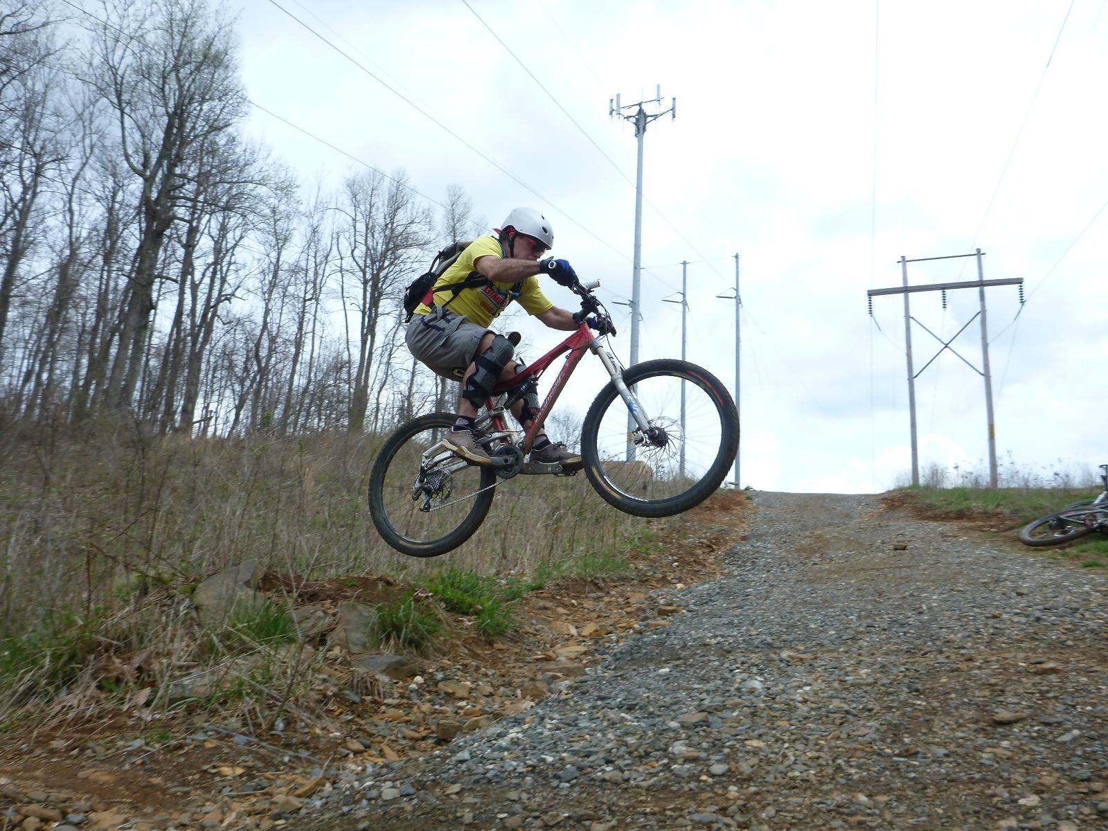 A mountain biker performing a jump on a gravel trail, with a backdrop of leafless trees and power lines under a cloudy sky. Rocky Knob Park mountain bike trail.