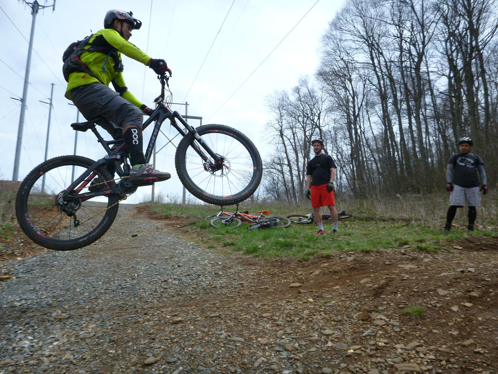 A mountain biker performing a jump on a trail, while two spectators watch nearby. The biker is wearing a bright green jacket and a helmet, and the surrounding area features trees and power lines in the background. Two other bicycles are lying on the ground, and the trail is rocky with patches of grass. Rocky Knob Park mountain bike trail.